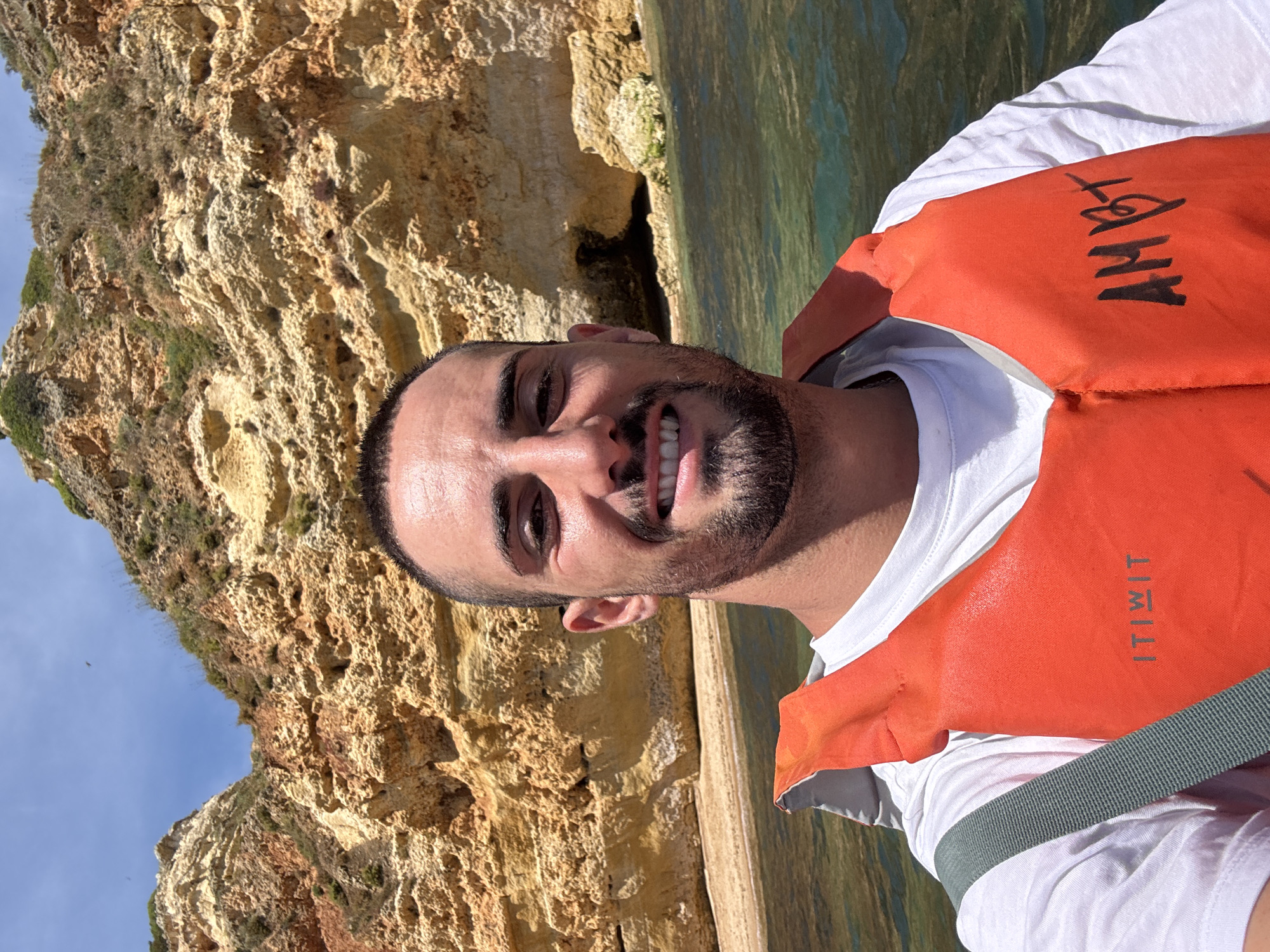 The image features a man taking a selfie outdoors, positioned in a body of clear water adjacent to impressive rocky cliffs. The man, smiling broadly and looking directly at the camera, has short dark hair and a neatly trimmed beard. He is wearing a white t-shirt underneath a bright orange life jacket.
The background is dominated by a rugged, light-brown rocky coastline or sea cliffs, showing textured surfaces and some sparse green vegetation. Below the cliffs, the water is remarkably clear, reflecting the sunlight and revealing parts of the seafloor. In the mid-ground, a dark opening, possibly a grotto or cave entrance, is visible at the base of the cliffs where the water meets the rock. A patch of clear blue sky is visible in the upper left of the frame.
The man's life jacket displays visible text. The brand name "ITIWIT" is printed vertically on the right side of the jacket, and a handwritten or drawn text, which appears to read "AMBY" or "AMBT," is present in black marker on the upper left side. The bright, direct sunlight suggests it is daytime, likely mid-morning or afternoon, and the clear sky indicates good weather. The overall scene depicts a person enjoying a water-based activity, such as kayaking or boating, in a scenic coastal environment.