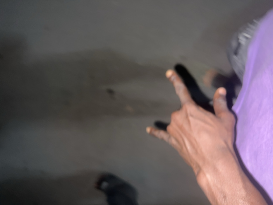 An image captured in Birnin Kebbi, Nigeria, depicts a human left hand in the foreground making the "rock on" gesture (index and pinky fingers extended, middle and ring fingers curled, thumb extended). The hand exhibits dark skin tone and is partially obscured by a light purple garment at the wrist. Directly behind and below the hand, two distinct dark shadows are cast on a light grey-brown, possibly concrete or asphalt, ground surface. These shadows are elongated and appear to be cast by human figures, with one shadow suggesting a torso and leg outline. The environment is dimly lit, and the ground surface shows minor discolored marks. No other subjects or objects are distinctly identifiable.
