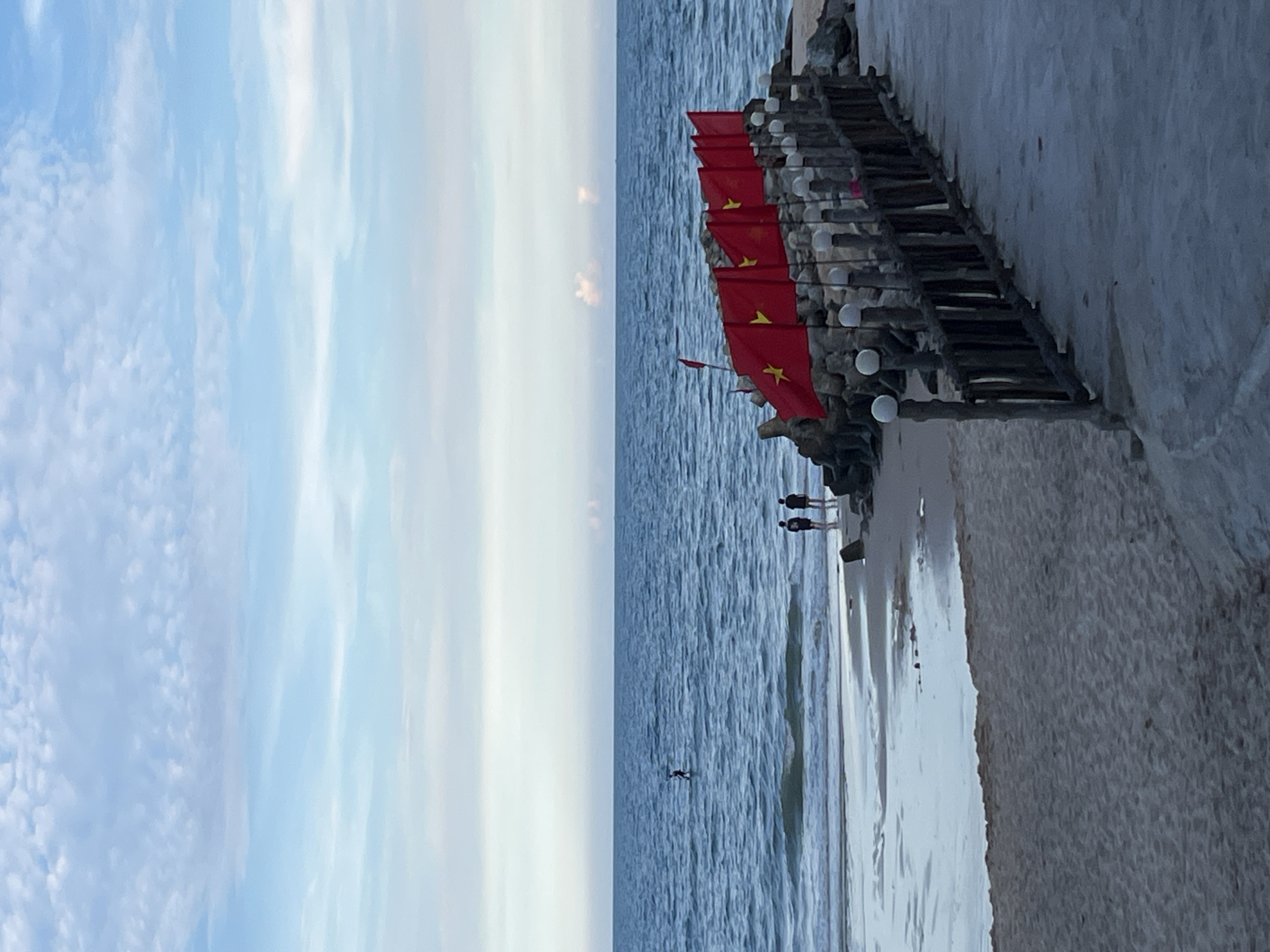 The image depicts a tranquil beach scene in Phu Thuy Ward, Vietnam, under a partially cloudy sky. The ocean is a deep blue, with gentle waves lapping at the sandy shore. In the foreground, a paved walkway leads to a rustic wooden fence adorned with spherical lights. A line of vibrant red flags, each featuring a yellow star, is mounted along the fence, creating a festive atmosphere.
Further down the beach, two individuals are silhouetted against the water, standing near the shoreline. In the distance, another person is visible on a paddleboard in the ocean. The sand itself is light brown, marked by the receding tide, which has left behind shimmering pools of water. The overall impression is one of peaceful coexistence between nature and human activity.