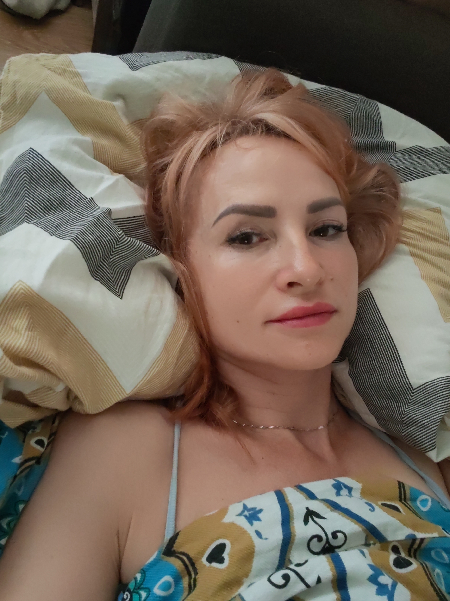 A woman with reddish-blonde hair lies in bed, looking at the camera. She is wearing a light blue spaghetti strap top and a silver necklace. The bed is covered with a patterned duvet and pillows with geometric designs. The background is blurred, showing what appears to be a dark couch and a wooden floor. The image does not contain any information that would allow for a specific location within the city of Chaikovsky, Russia.