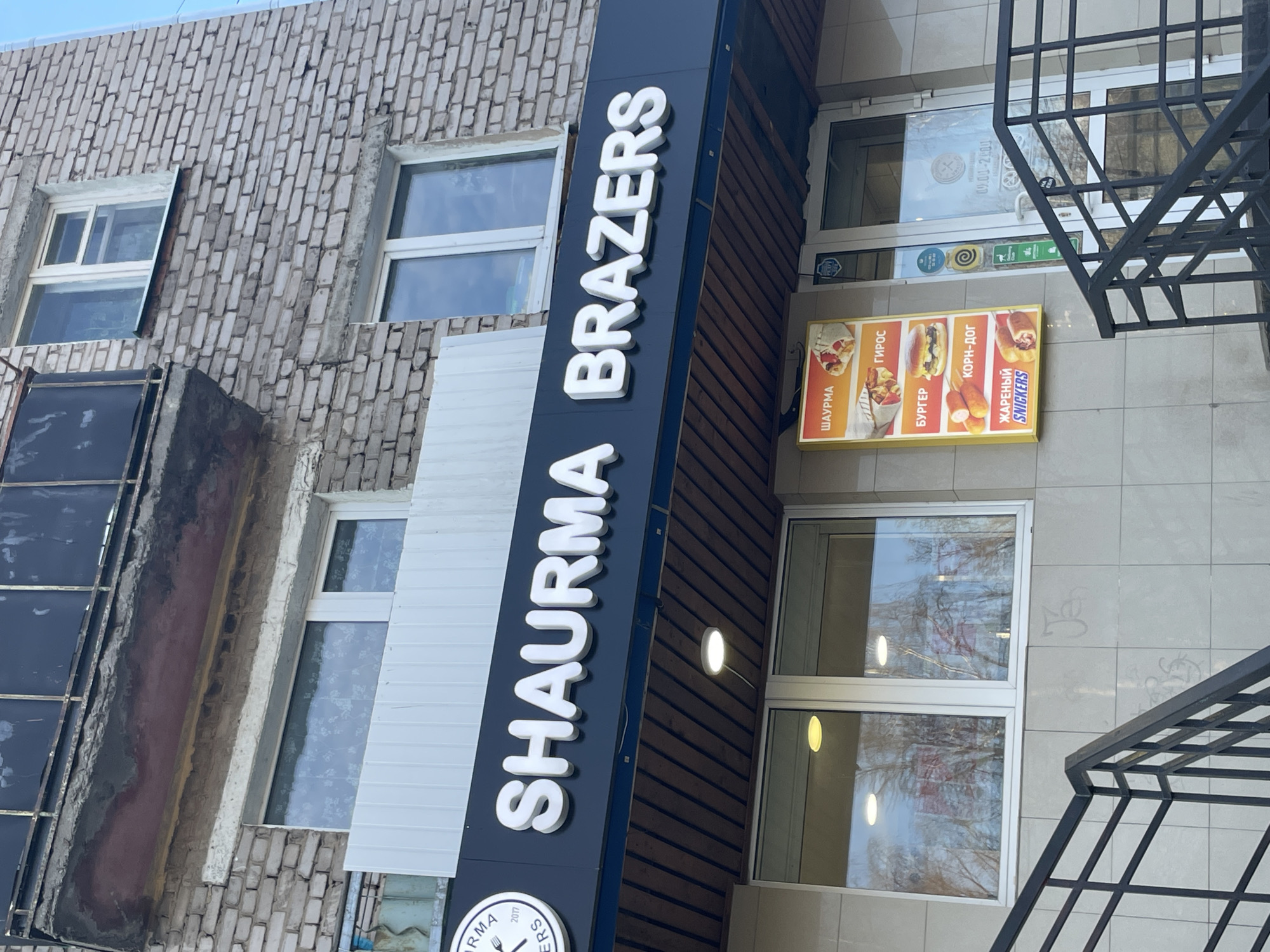 This media file captures the exterior of a fast-food establishment named "SHAURMA BRAZERS" located on the ground floor of a multi-story building, likely in an urban setting. The building features a light-colored, somewhat aged brick facade with several windows visible on the upper floors.
A prominent vertical sign with white, raised letters against a dark background displays "SHAURMA BRAZERS". Below the main text, a circular logo can be partially seen, which includes a fork and knife symbol and the year "2017". To the right of this main sign, an illuminated menu board showcases various food items. The items listed, primarily in Cyrillic, are "ШАУРМА" (Shawarma), "ГИРОС" (Gyros), "БУРГЕР" (Burger), "КОРН-ДОГ" (Corn-Dog), and "ЖАРЕНЫЙ SNICKERS" (Fried Snickers).
The ground floor features large glass windows and an entrance door, reflecting the outdoor environment, including what appears to be trees or distant buildings. A black metal railing, possibly part of an outdoor staircase, is visible on the right side of the image, extending upwards. The overall scene appears to be taking place during the daytime, under natural light, though the exact time of day or weather conditions (e.g., sunny or overcast) are not definitively clear. The use of Cyrillic text aligns with the provided location context of Izhevsk, Russia. No people or active events are directly visible in the frame.