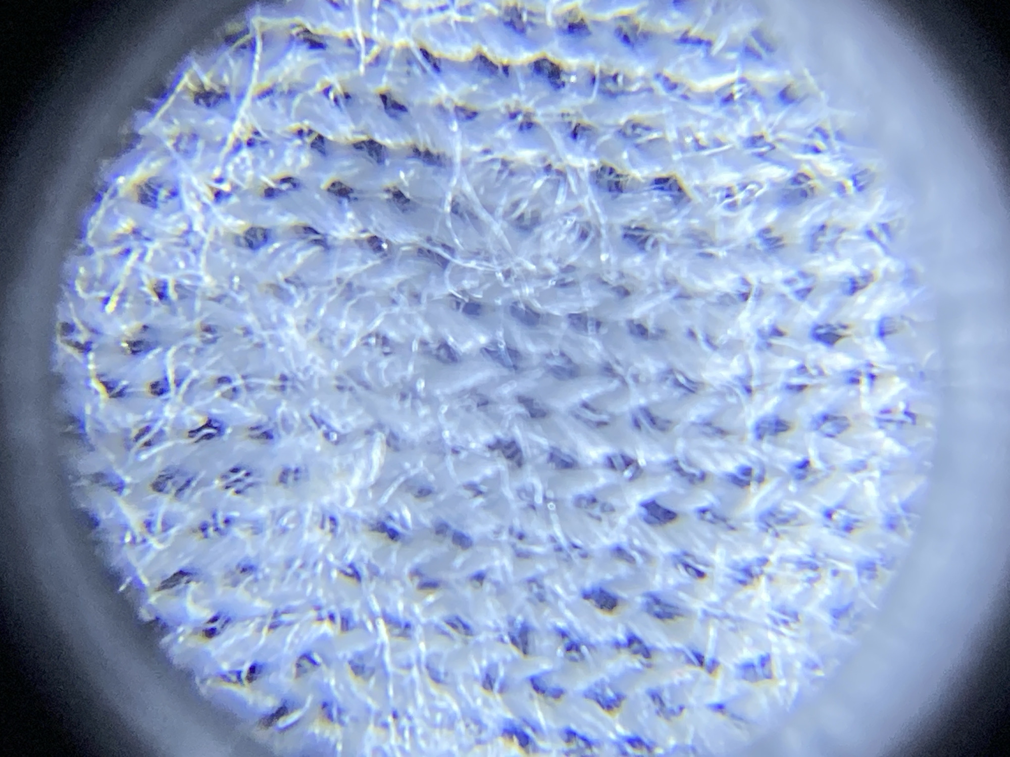 The image is a close-up, likely microscopic, view of a white, woven fabric. The weave appears to be a pattern of interlocking loops or threads, creating a textured surface with small openings. Individual fibers are visible, appearing somewhat fuzzy and irregular. The lighting casts shadows and highlights, emphasizing the three-dimensional structure of the material. The image is circular with a dark vignette around the edges, suggesting it was taken through a lens or optical instrument. There is no indication of a city, location, people, or any actions occurring. The image solely focuses on the texture and structure of the fabric.