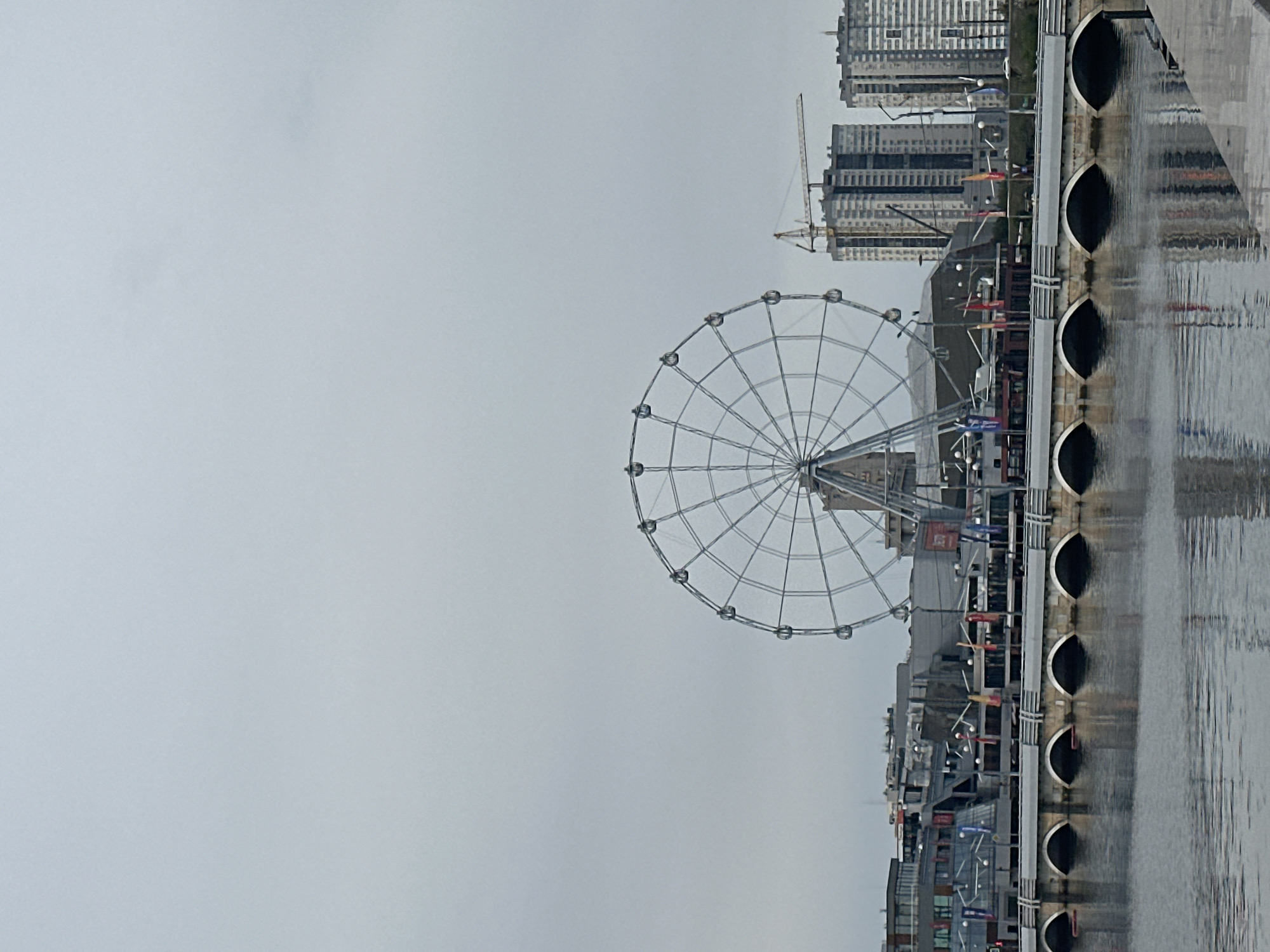 The image shows a Ferris wheel and several buildings along a waterfront in an urban environment under an overcast sky. The Ferris wheel is a prominent feature in the center of the frame, with its metal spokes and gondolas clearly visible. To the right of the Ferris wheel are several modern high-rise buildings under construction, indicated by a construction crane. In the foreground, a body of water reflects the scene. A bridge with arched supports spans across the water, connecting the waterfront to another area. Buildings and structures are visible on the waterfront, with flags displayed along the bridge and some of the buildings. The overall atmosphere is somewhat subdued due to the cloudy weather.