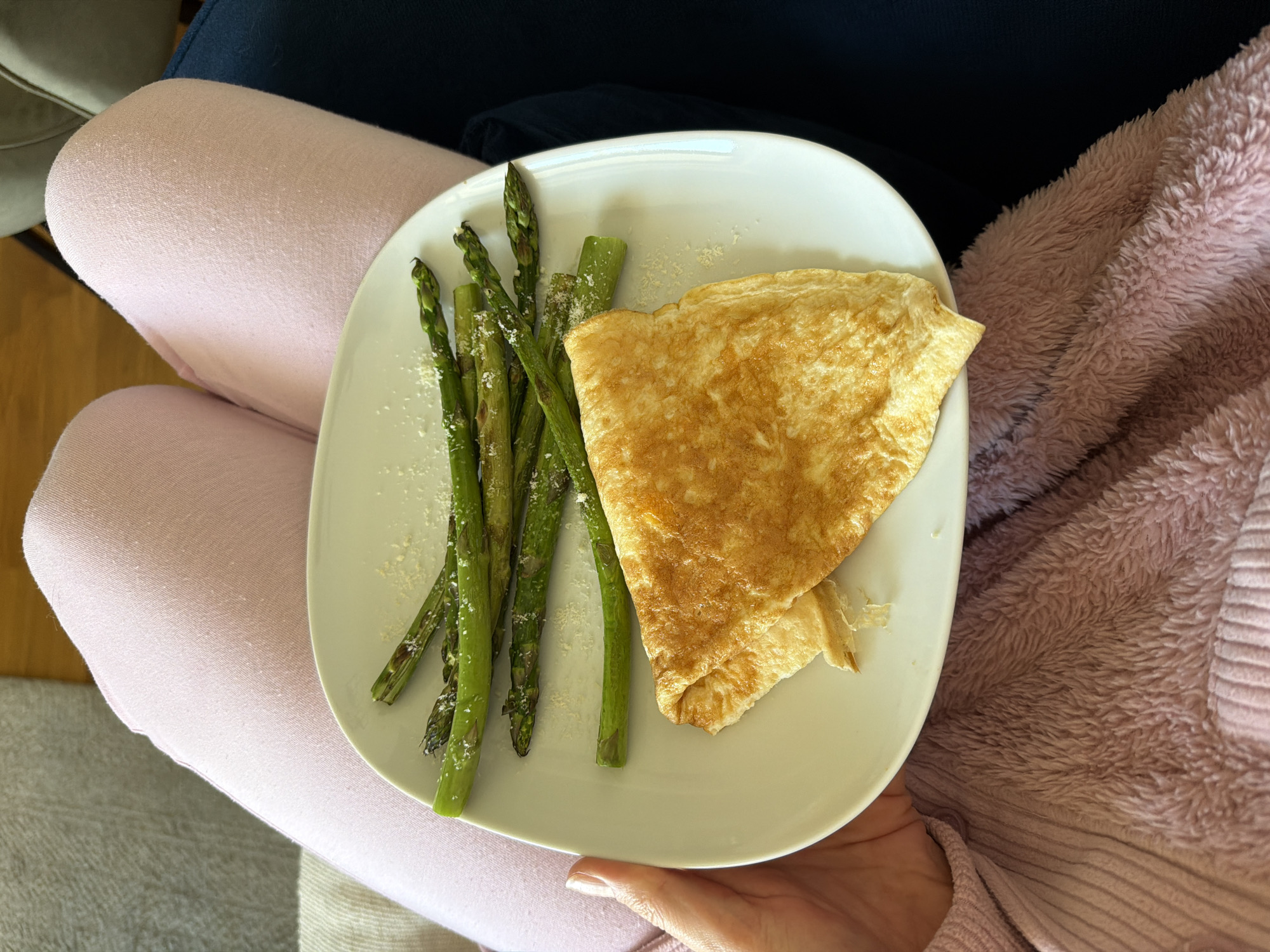 A plate with an omelet and cooked asparagus rests on a person's lap. The individual is wearing pink leggings on their left leg and a fluffy pink sweater on the right. Some white particles are sprinkled on the asparagus. The background shows a hint of a dark blue garment and a beige-toned surface. The location is not discernible from this image, and it is not possible to determine if the image was taken in Belgrade, Serbia.