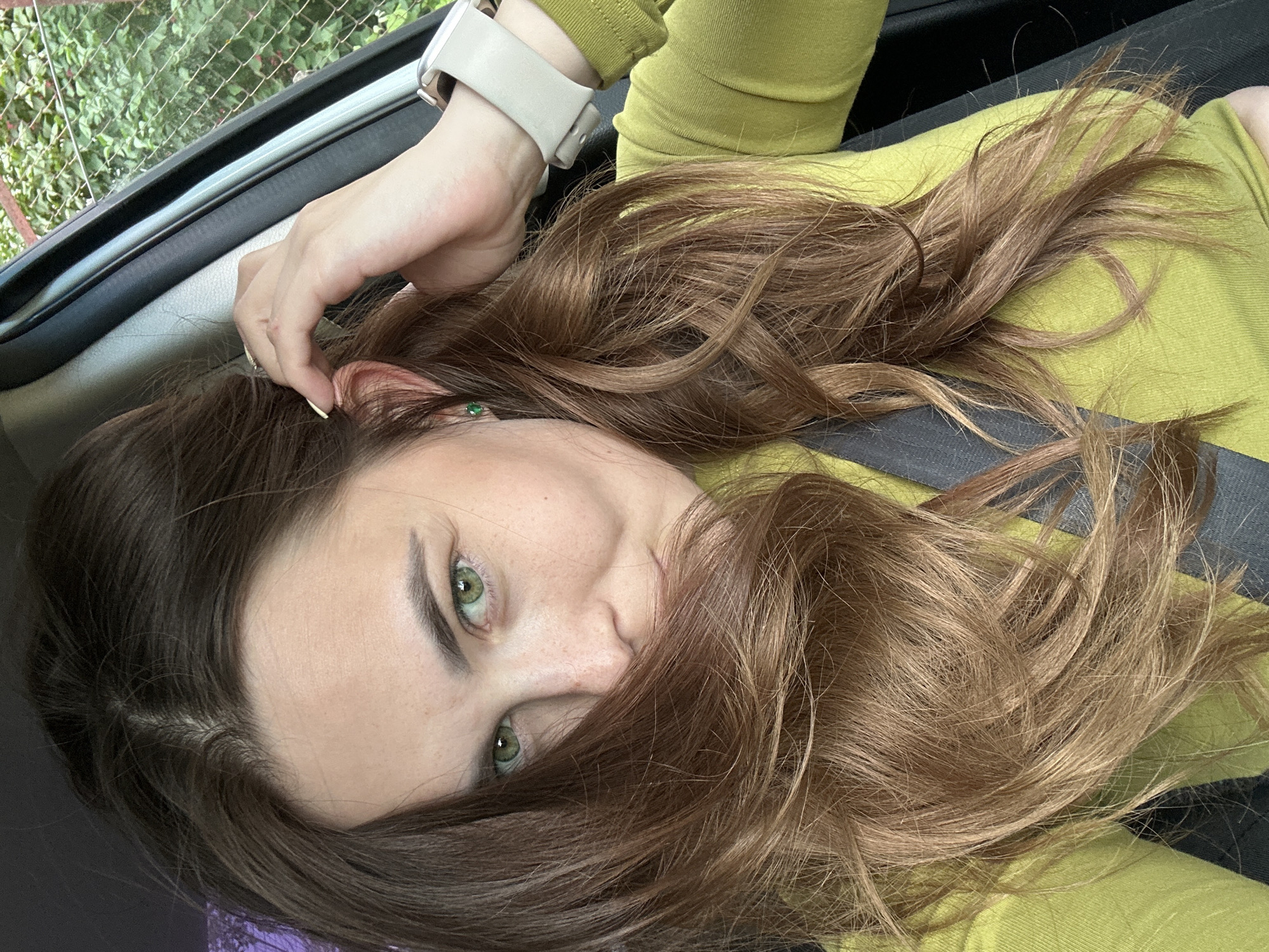 A woman with brown hair and green eyes is sitting in a car. She is wearing a lime green long-sleeved shirt and a grey seatbelt. Her hair is partially covering her face, and she is touching her hair with her right hand. Her left hand is resting on her lap. She is wearing a watch on her left wrist and an earring in her left ear. The car interior is visible, including the window and car door. Outside the car, greenery and a chain-link fence are visible. The location is identified as Ilyinsky, Russia.