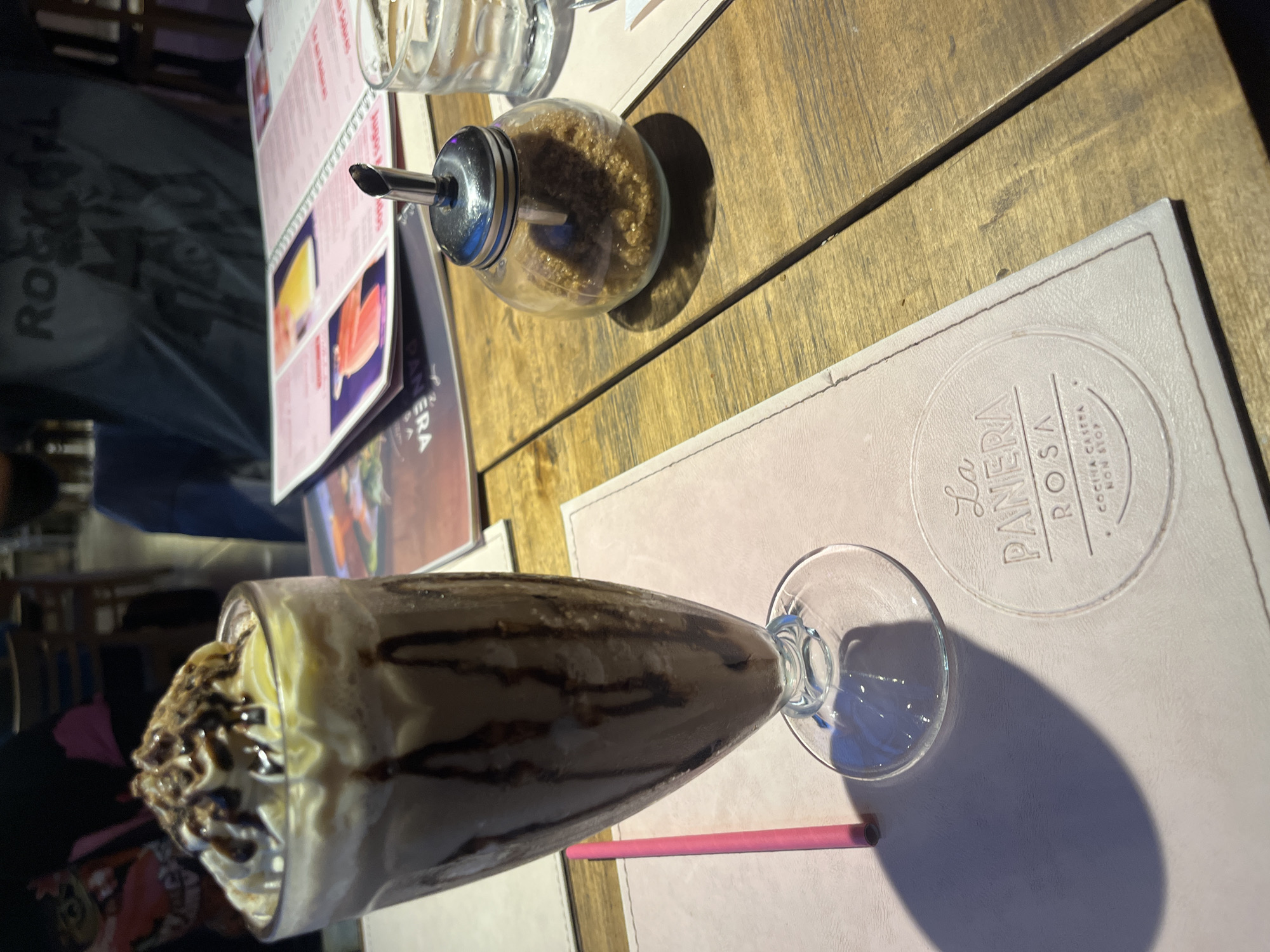 The image depicts a table setting at "La Panera Rosa" restaurant in Lomas de Zamora, Argentina. A chocolate milkshake, topped with whipped cream and chocolate drizzle, sits on a pink placemat. A pink straw rests beside the milkshake glass. A sugar dispenser filled with brown sugar and a glass are positioned on the wooden table, along with a menu. A person wearing a "Rock" t-shirt is partially visible in the background.