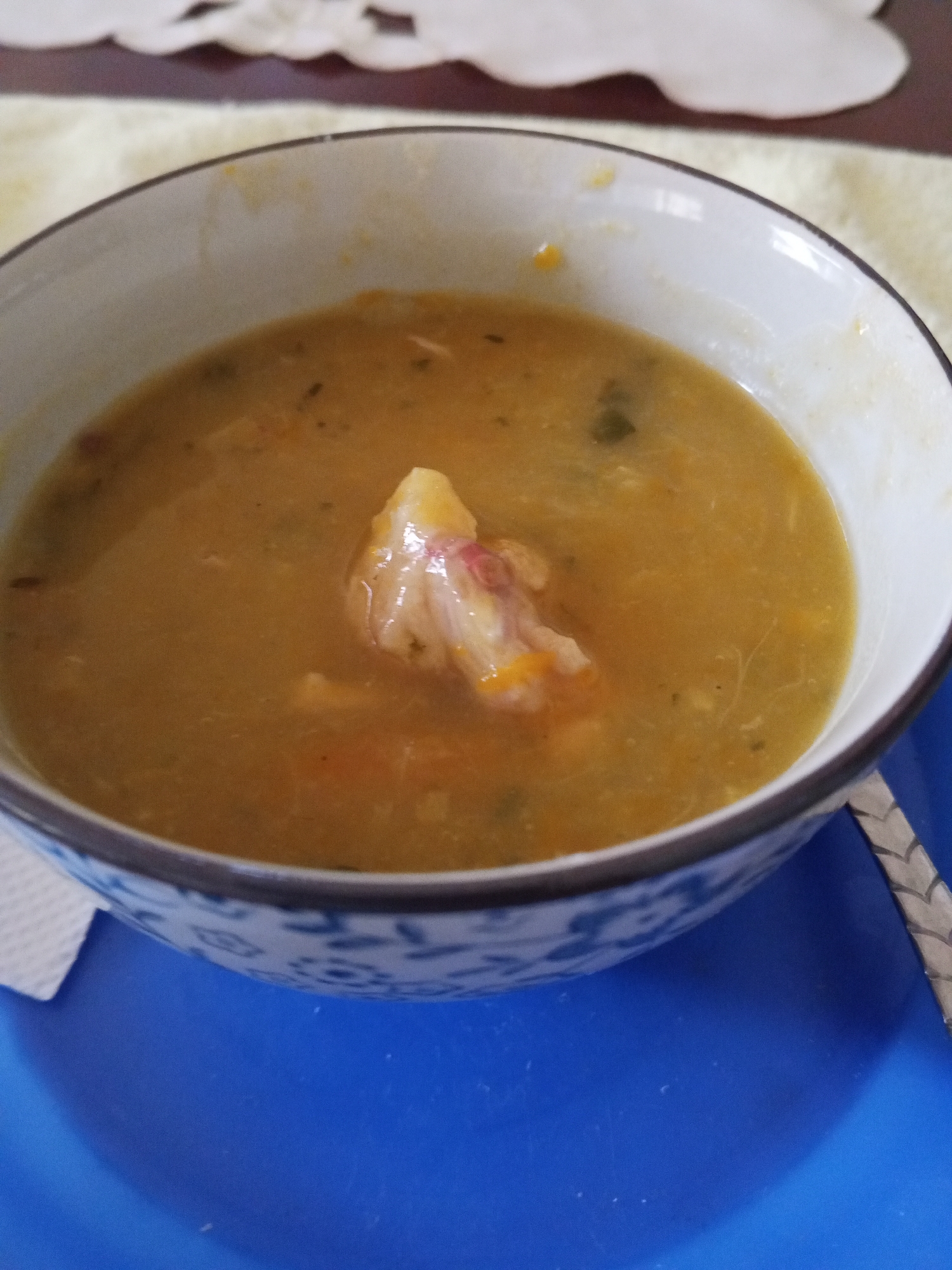 A bowl of soup sits on a blue plate. The soup appears yellow with visible ingredients, including a large piece of what seems to be meat or vegetable. The bowl itself has a white interior with a blue decorative pattern on the exterior. A white napkin is to the side of the bowl and a utensil is to the right. A beige tablecloth is visible in the background.
Based on the question, this image was taken in Tinaquillo, Venezuela.