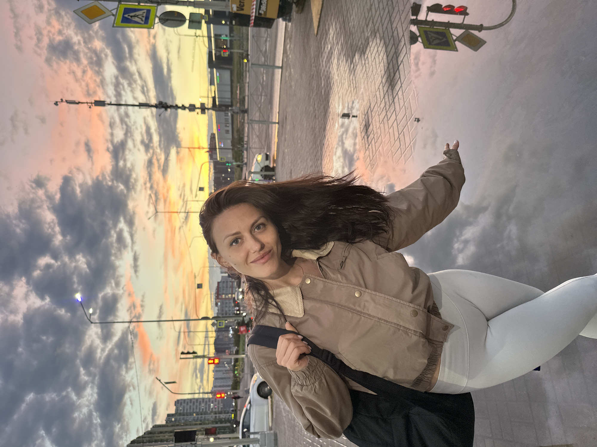 A woman with dark hair and brown eyes, wearing a beige jacket and white leggings, poses for a photo on a wet sidewalk at dusk. Her body is angled towards the camera, and her head is tilted to the left. She holds a black bag over her left shoulder. The background shows a city street with buildings, traffic lights, and streetlights. The sky is cloudy with hints of orange and pink from the setting sun. The wet pavement reflects the sky and the surrounding environment.
