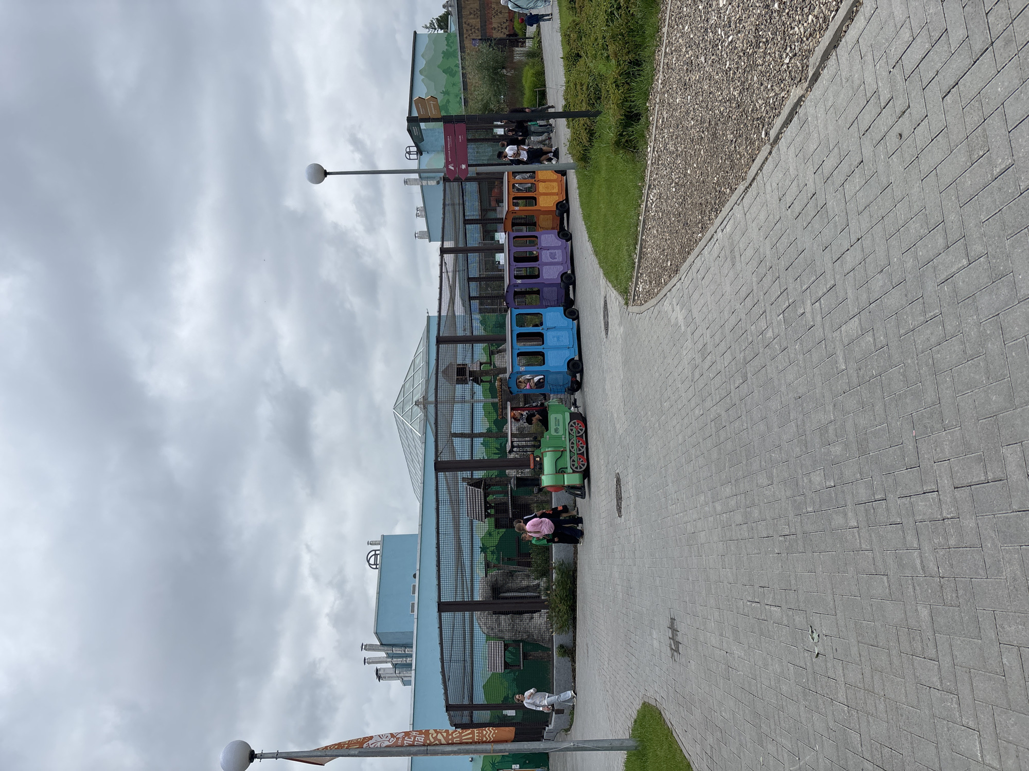 The image captures an outdoor scene at the Perm Zoo in Perm, Russia, on a cloudy day. In the foreground, a paved walkway curves to the right, bordered by a gravel path and lush green grass. A lamppost with a spherical light fixture stands on the left side of the walkway.
Further back, a series of colorful, child-sized train cars—blue, purple, and orange—are parked on a track. A green locomotive is attached to the front of the blue car. A few people, including children and adults, are visible near the train, suggesting a recreational area within the zoo.
Behind the train, a large enclosure with a mesh fence encloses a section of the zoo. Within the enclosure, there is a rocky structure, trees, and small wooden structures, resembling animal habitats. A building with a light blue facade and industrial-looking chimneys is situated behind the enclosure, with a domed glass structure visible on its roof. The background features a mural of a green, hilly landscape with trees painted on a wall.
Directional signs are also present near the train, indicating different areas within the zoo. The overall atmosphere suggests a family-friendly attraction within the zoo premises.