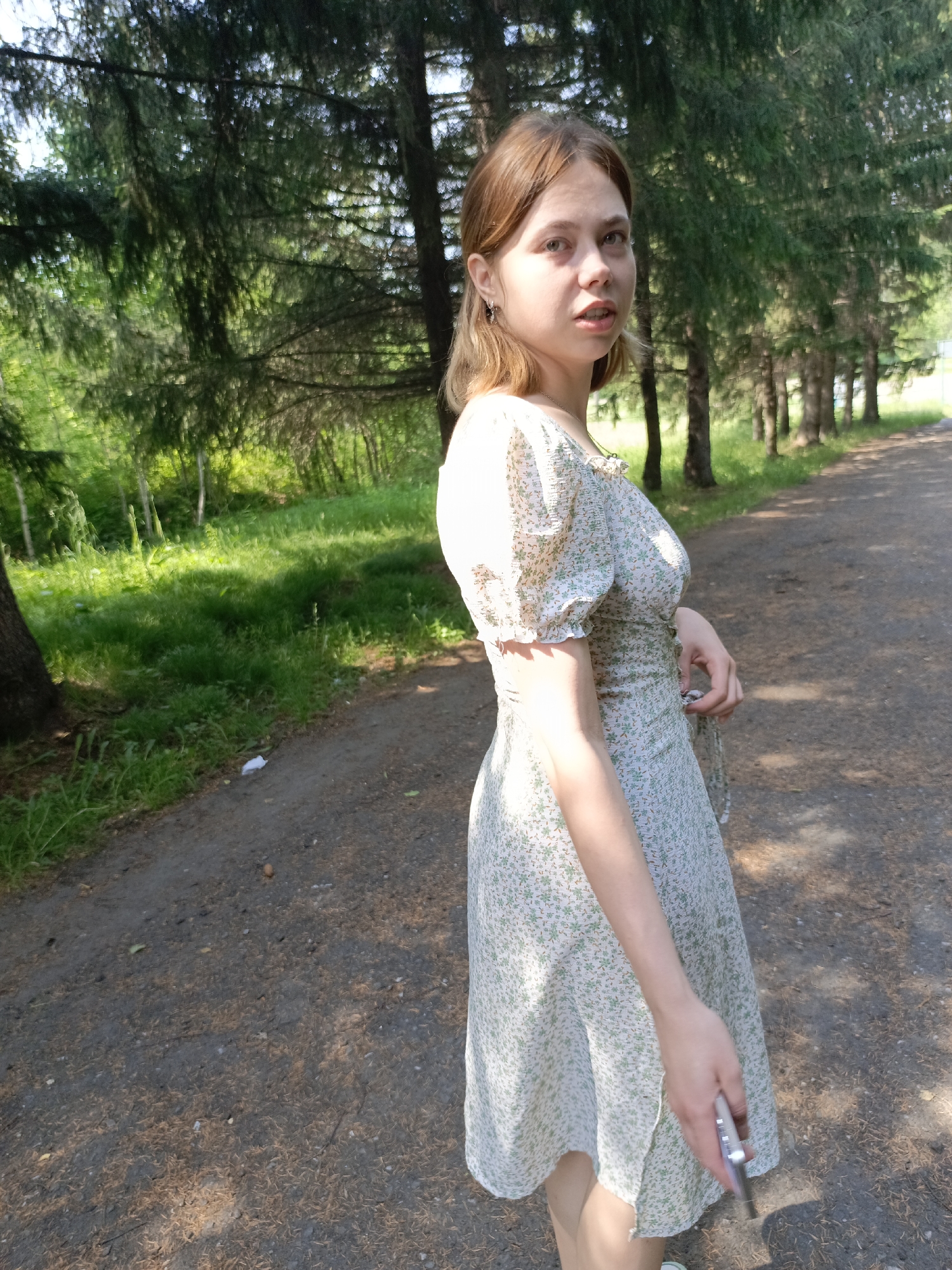 In Zalesovo, Russia, a young woman with light brown hair stands on a dirt path lined with evergreen trees. She is wearing a light-colored floral dress and holding a silver object in her right hand. Sunlight filters through the trees, casting dappled shadows on the path and the surrounding green grass.