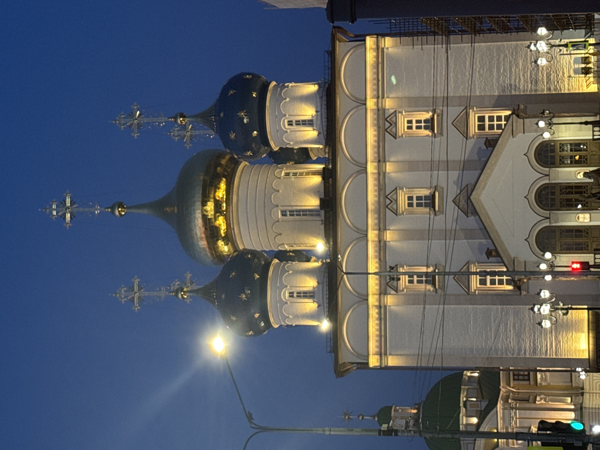 The image shows the illuminated domes and towers of a Russian Orthodox church or cathedral at night, likely located in the city of Tver, Russia, given the style of architecture. Three prominent domes with crosses on top are visible against a blue sky, one gold and two dark blue adorned with stars. To the right of the domes is a multi-storied white building with windows and arched features, illuminated with golden light. Streetlights are present and there are power lines that are crossing in front of the image. A traffic signal is present, showing a green light.