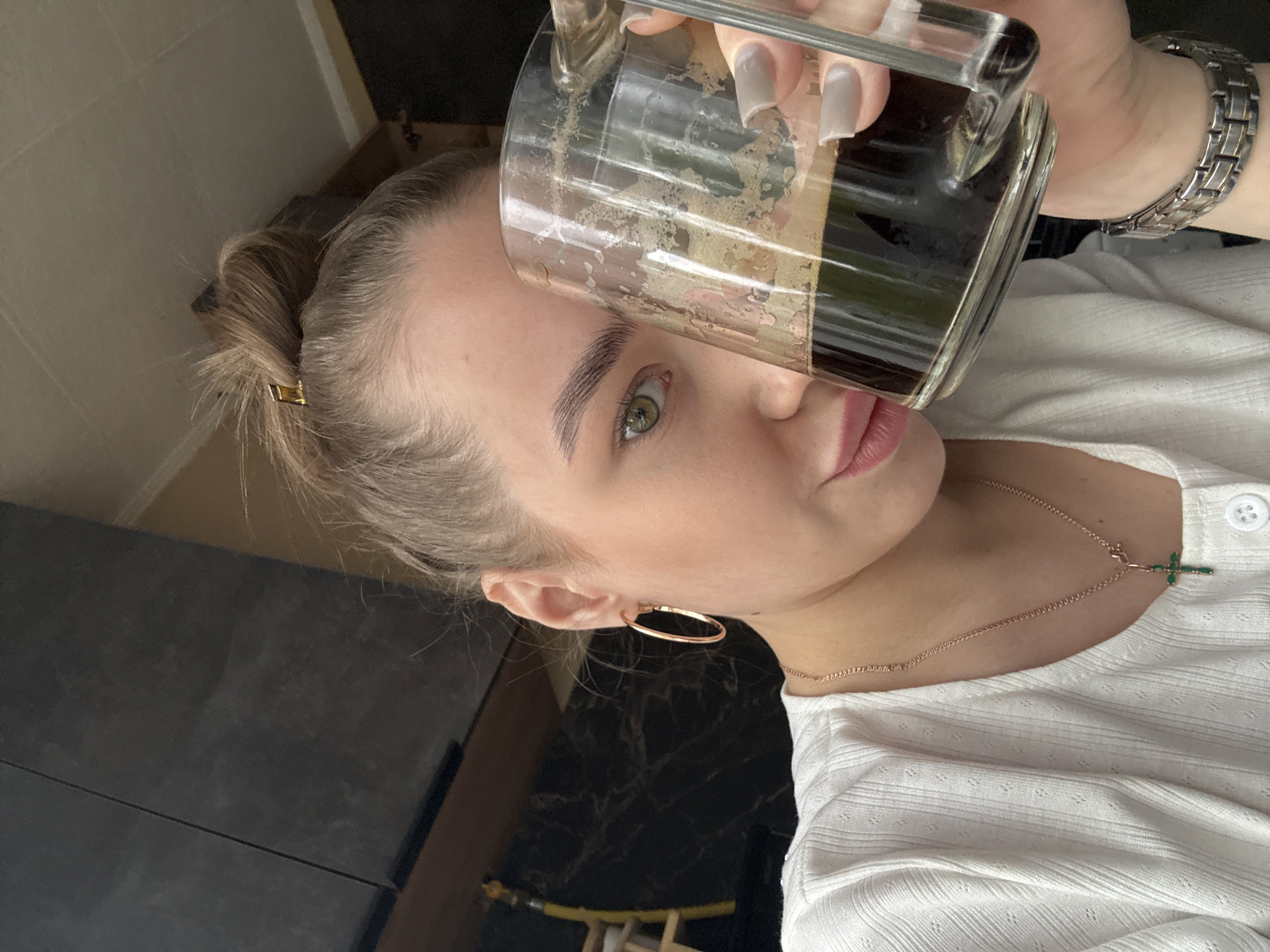 A young woman with blonde hair pulled back in a bun is holding a clear glass mug filled with a dark liquid, likely coffee or tea, close to her mouth. She has light skin, green eyes, and manicured nails with a light gray polish. She is wearing gold hoop earrings, a delicate gold necklace with a small green cross pendant, and a white textured shirt with a button at the collar. A silver watch is visible on her wrist. The background appears to be an interior space with dark gray cabinets and a glimpse of a wooden surface. The location in the city of Chaikovsky, Russia, is not determinable from the provided visual information.