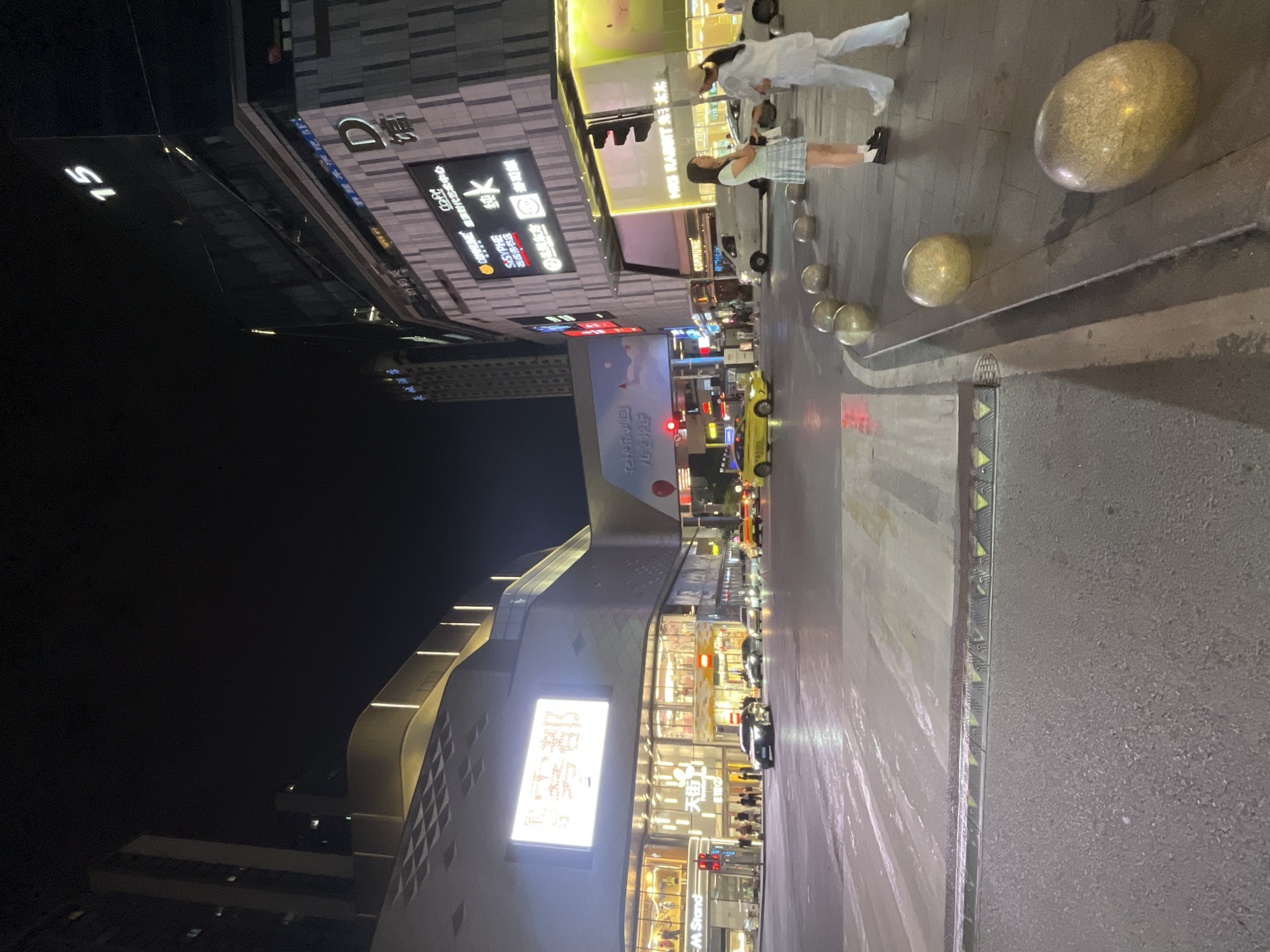 The image captures a nighttime scene at an intersection in Yuzhong District, China, dominated by illuminated buildings and streetlights. A large modern building with a prominent "D" logo and various Chinese characters and signage is visible on the right. Across the street, another multi-story building features a large, bright screen displaying text in Chinese characters and a stylized cartoon image. The intersection is paved and shows signs of recent rain, with wet reflections on the road surface. Several vehicles, including a black sedan, a white van, and yellow taxis, are present at the intersection. Two young women, one in a green and white checkered skirt and the other in white clothing, stand on the sidewalk near a traffic light. Decorative golden, egg-shaped bollards are lined along the sidewalk. The sky is dark, indicating it is nighttime.