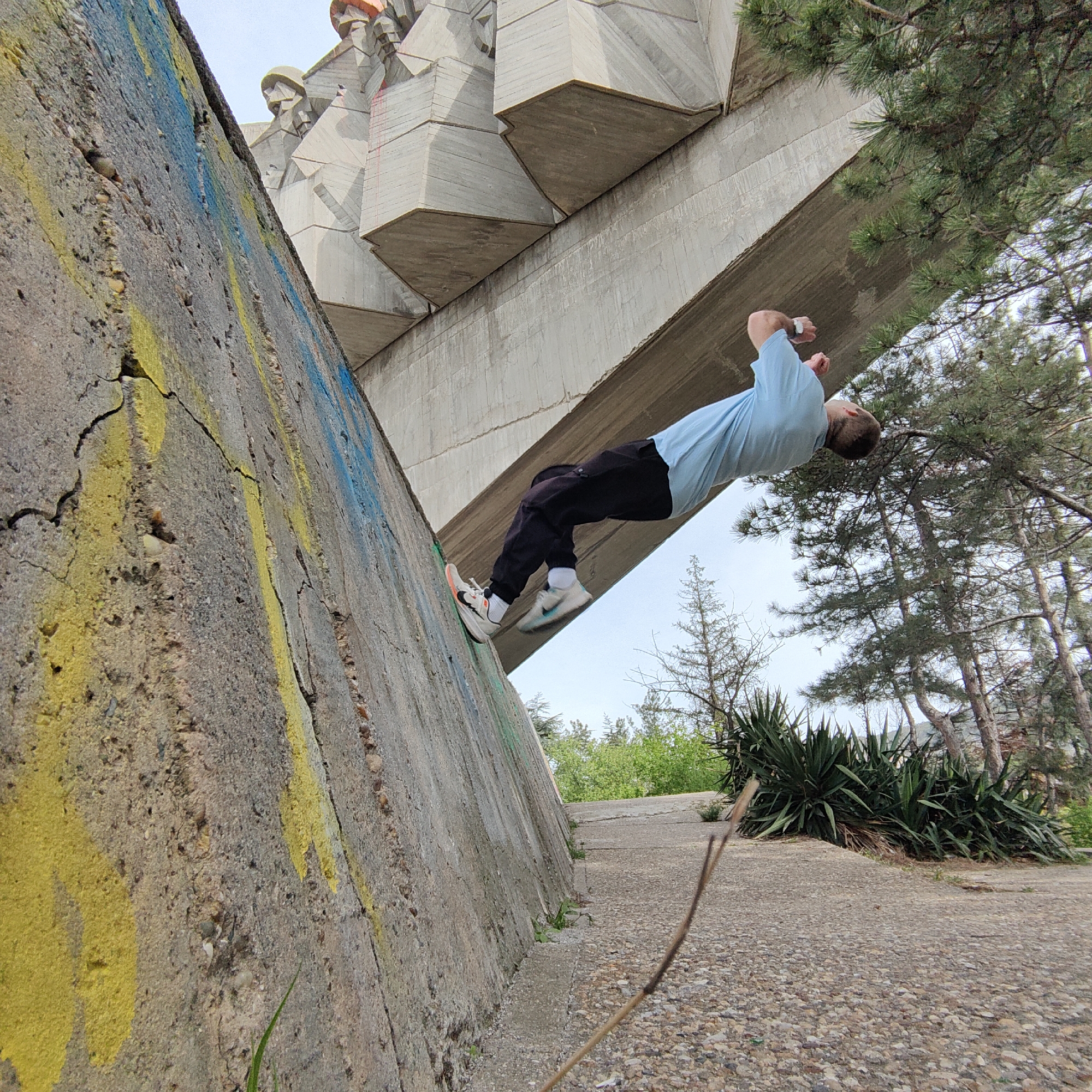 A man is captured mid-backflip in an outdoor setting near a large concrete structure, which is part of the Monument of the Soviet-Bulgarian Friendship in Varna, Bulgaria. The man, wearing a light blue shirt, dark pants, and sneakers, is positioned with his feet against a graffiti-marked concrete wall. The structure overhead consists of gray concrete segments, forming an overhang. In the background, there are trees and vegetation. The ground is a paved surface with pebbles. A small branch lies in the foreground. The sky is partly visible above the structure.