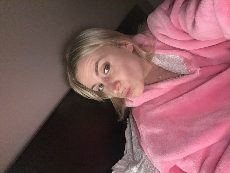 A fair-skinned woman with blonde hair poses with pursed lips. She is wrapped in a fluffy pink robe. A small silver necklace is visible at her neck. She appears to be lying in bed, indicated by patterned bedding below. The background consists of a dark surface, possibly a headboard or wall. The image is slightly angled, giving a tilted perspective.
