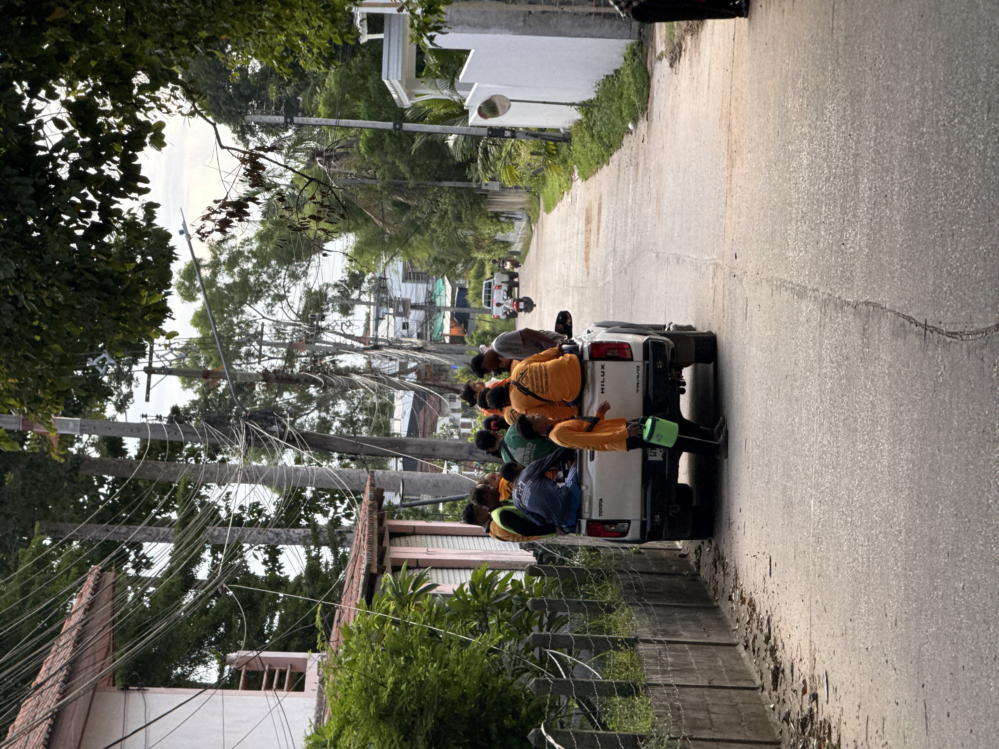In the city of Ko Samui District, Thailand, a group of individuals are seen riding in the back of a white Toyota Hilux pickup truck. The truck is parked on a paved road lined with a concrete fence and lush green trees. Several people, mostly wearing orange shirts, are seated and standing in the truck bed, some appear to be workers. One person, also in an orange shirt, is walking beside the truck, carrying a green bucket. In the background, motorcycles and a white SUV are visible further down the road, suggesting activity and traffic. Overhead, a dense network of power lines crisscrosses the scene, indicative of urban infrastructure. The overall impression is a snapshot of daily life and transport in a tropical setting.