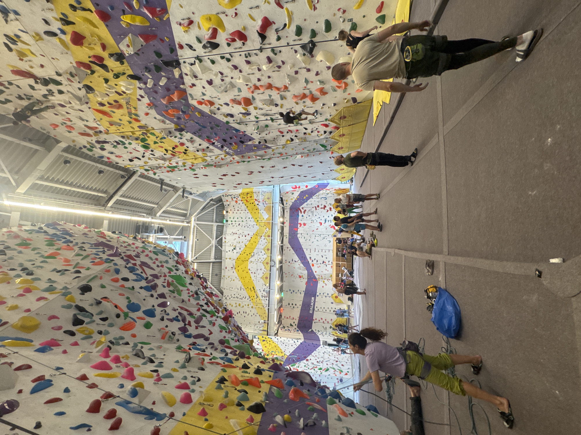 This image captures a vibrant indoor rock climbing gym, likely in Sofia, Bulgaria, filled with climbers and colorful equipment. The facility features numerous climbing walls of various inclines and heights, densely adorned with a multitude of brightly colored holds in red, yellow, blue, green, orange, purple, black, white, and pink. Sections of the white walls are highlighted with distinct vertical stripes in yellow and purple. The word "BALKAN" is prominently visible vertically on one of the purple-striped climbing walls.
Several individuals are engaged in climbing. One person in a purple shirt and green shorts, wearing a harness and climbing shoes, is actively ascending a wall in the foreground on the right. Higher up and to the right, another climber in a light shirt and dark pants scales an overhanging section. Other climbers are visible on different walls in the background, showcasing various levels of ascent. On the grey floor to the right, a group of people stand, some appearing to observe or prepare for their turn. A blue bag rests on the floor near a climber.
The gym is brightly lit by artificial overhead lighting, revealing an industrial-style ceiling with metallic beams and corrugated panels. The atmosphere suggests an energetic and focused recreational activity, likely taking place during active hours of the day or early evening.