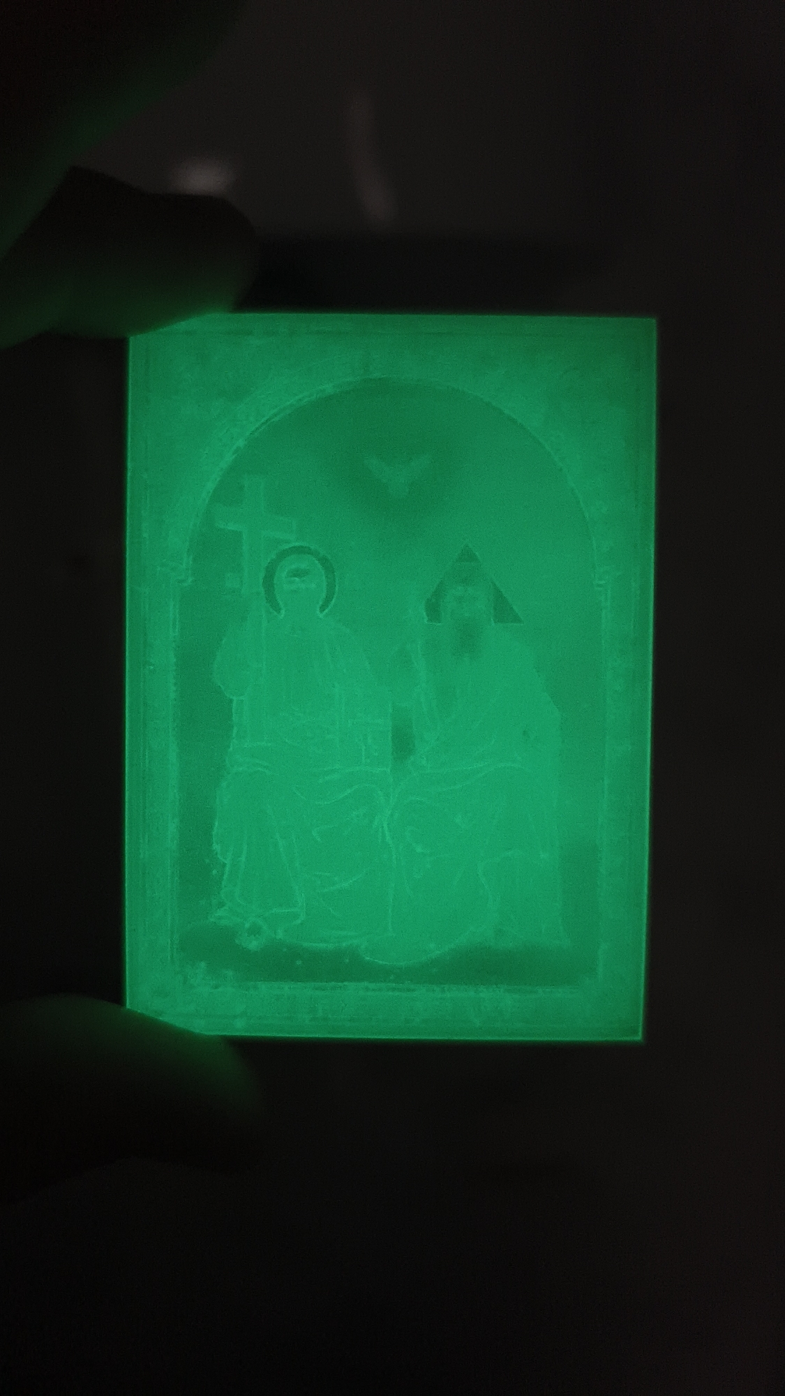 An image displays a rectangular, translucent object glowing with a bright green phosphorescence in a dark environment. The object appears to be a religious icon or amulet, etched with the figures of two individuals. The figure on the left is depicted with a halo and is holding a cross. The figure on the right has a triangular headdress and appears to be seated next to the first figure. Above the figures, an arch frames them, and the faint outline of a bird or dove is visible. The object is held between the fingers of a hand, suggesting it is small and portable. The background is entirely black, indicating a low-light or nighttime setting. The scene is likely a close-up shot intended to highlight the glowing quality of the object.