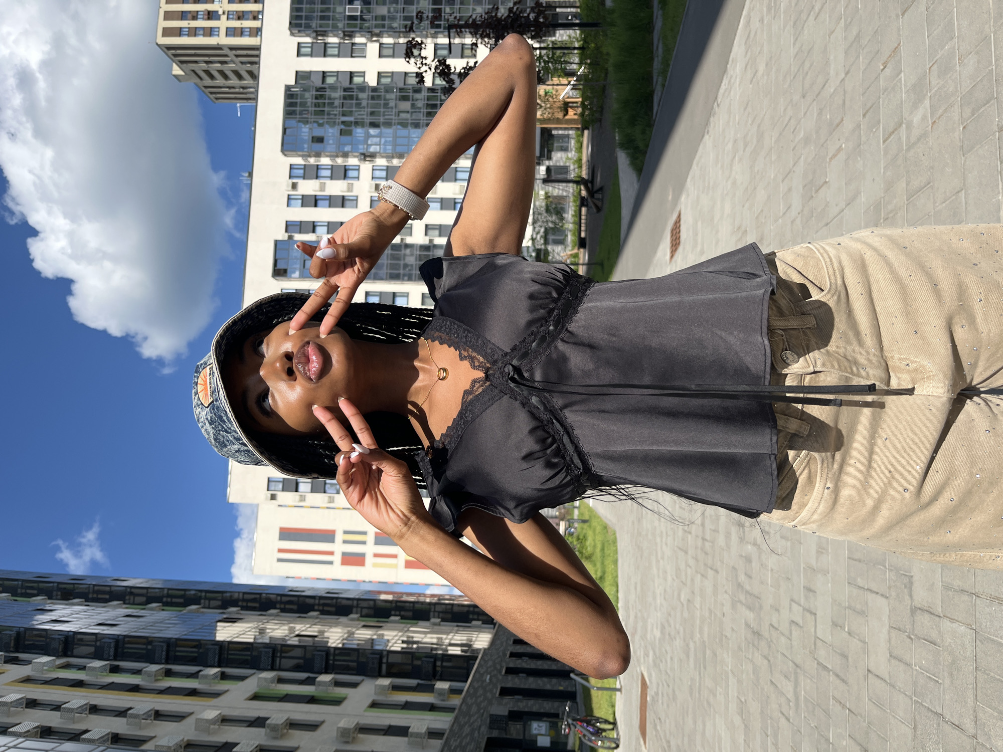 A young Black woman is posing for a photo outdoors in what appears to be a modern urban setting. She is wearing a black, sleeveless top with lace trim and a v-neck, paired with tan pants adorned with small rhinestones. A patterned bucket hat covers her head, and she is accessorized with a gold necklace and a beaded bracelet. Her fingers are positioned in a peace sign gesture near her face, and her lips are puckered. The background features contemporary apartment buildings with large windows and colorful architectural elements, suggesting a relatively new development. Paved walkways and patches of green grass indicate a public outdoor space. The lighting suggests it is daytime with a clear sky and some clouds. The presence of the apartment buildings and the overall modern aesthetic are consistent with areas found in Kazan, Russia.
