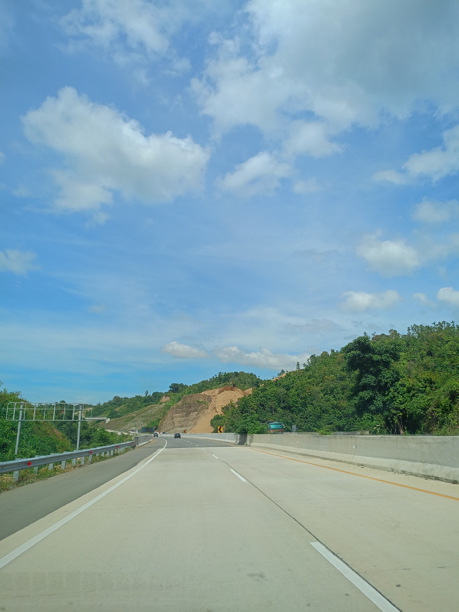 The image captures a view from what appears to be the driver's seat of a vehicle traveling along a multi-lane highway near Suyo Paloh, Indonesia. The road is concrete, marked with white lines separating lanes, and a yellow line indicating the edge of the road on the right. A grey crash barrier runs along the left side of the road, with a metal guard rail above it. In the distance, other vehicles are visible on the road. The sky is a light blue with scattered white clouds. A hillside covered with lush green vegetation is present to the right of the road. A section of exposed earth is visible in the hillside. Signage is visible along the roadside.