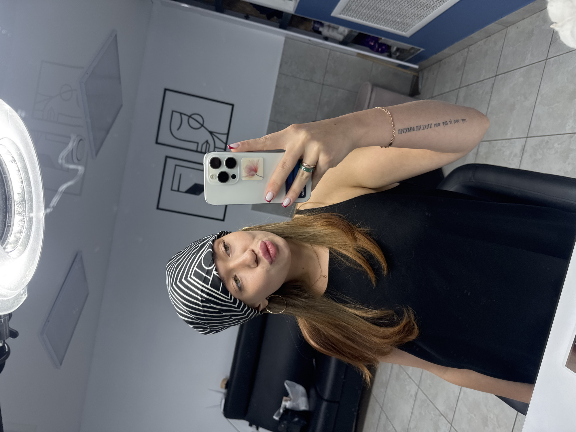 The image is a vertical, full-body mirror selfie taken by a young woman. She is wearing a black sleeveless top and a black and white striped bandana. She has long, light brown hair and is wearing large hoop earrings and a delicate bracelet on her left arm. She is also wearing two rings on her right hand, one of which appears to be green. Her nails are painted red and white.
In the background, there is a reflection in a mirror. The reflection shows two framed line drawings of faces, a bright ring light, and part of what looks like a salon or studio space with tiled floors and walls. On the woman's forearm, a tattoo is visible, consisting of Roman numerals and some other text that is difficult to discern. She is holding a white smartphone in her right hand, which is angled to capture the selfie. Her expression is playful, with her lips pursed. The lighting suggests it is daytime, and the indoor setting provides no weather cues. The overall impression is of a casual, self-aware moment captured for social media.