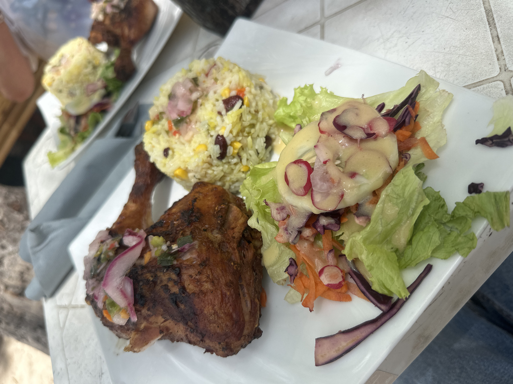 The image shows a plate of food, possibly taken at a restaurant in Le Gosier, France, given the setting. The plate features a piece of grilled or roasted chicken with a garnish of pickled onions and other vegetables. A portion of rice with corn and kidney beans is placed next to the chicken. A salad with lettuce, shredded carrots, sliced radishes, red cabbage, and a creamy dressing is also present on the plate. Another plate of food is partially visible in the background, suggesting that it is a shared meal setting. The table has a textured, possibly tiled, surface.