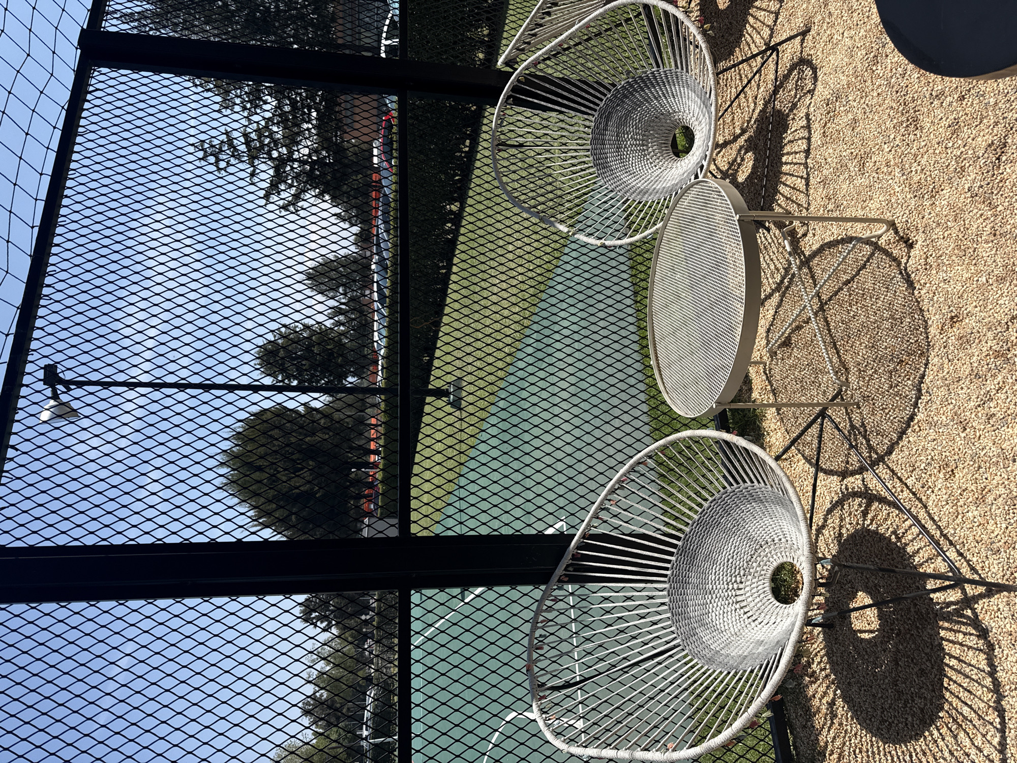 The image shows a seating area with two modern, white, basket-weave chairs and a small, round table on a tan gravel surface. The seating area is adjacent to a tennis or paddle court, visible through a black metal mesh fence. Trees and possibly other structures are visible beyond the court. The location is likely San Andrés Cholula, Mexico. The image is taken from an elevated perspective. Shadows from the chairs and table are cast on the gravel.