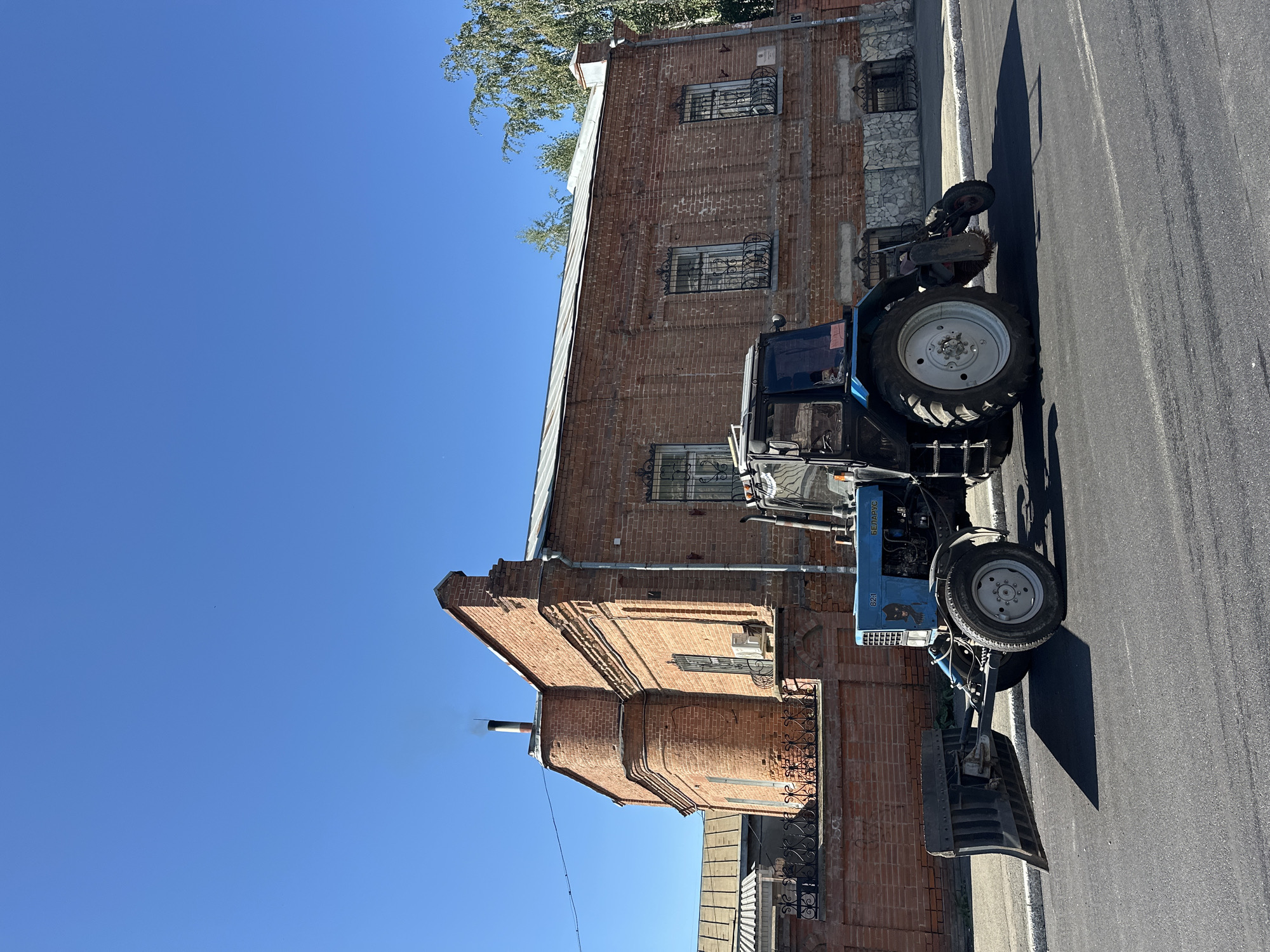The image shows a blue tractor with a snowplow attachment parked on an asphalt road in front of a brick building. The building has a metal roof, barred windows, and a chimney with smoke emitting from it. The sky is clear and blue. The tractor has the word "БЕЛАРУС" written on its side, which is a brand of Belarusian tractors. The scene is likely a street in Biysk, Russia, given the presence of a Belarusian tractor and the architectural style of the building. There are no people visible in the image. The tractor appears to be idle, and its snowplow is lowered to the ground. The environment suggests it could be a scene from a town or city where such equipment is used for maintenance.