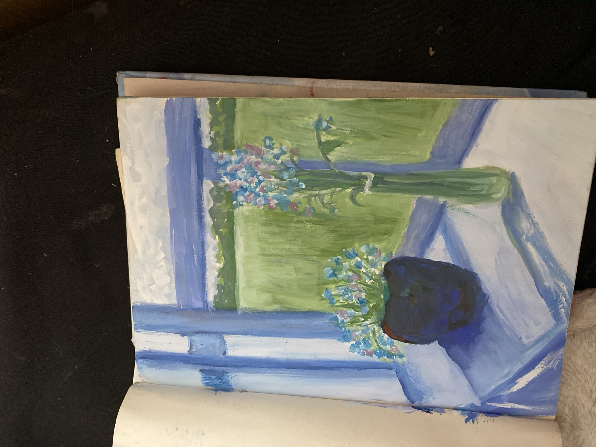 This is an image of a painting, likely in an art sketchbook, placed on a dark, textured surface. The painting depicts a still life of flowers, possibly forget-me-nots, with light blue and pink blossoms and green stems, arranged in a vase. The vase is dark blue and appears to be a rounded shape with some lighter blue tones. The background suggests an indoor setting looking out a window. There are prominent vertical and horizontal blue bands, suggesting a window frame and perhaps a windowsill. Beyond the window, a bright green, textured area is visible, indicative of grass or foliage, hinting at an outdoor scene under a bright sky. The overall color palette is dominated by blues, greens, and whites, creating a light and airy feel. The style of the painting is impressionistic, with visible brushstrokes and a focus on light and color rather than sharp detail. There is no discernible text or specific identifiable landmarks. The lighting suggests it could be daytime, with natural light coming from the depicted window. The emotional tone is serene and peaceful.