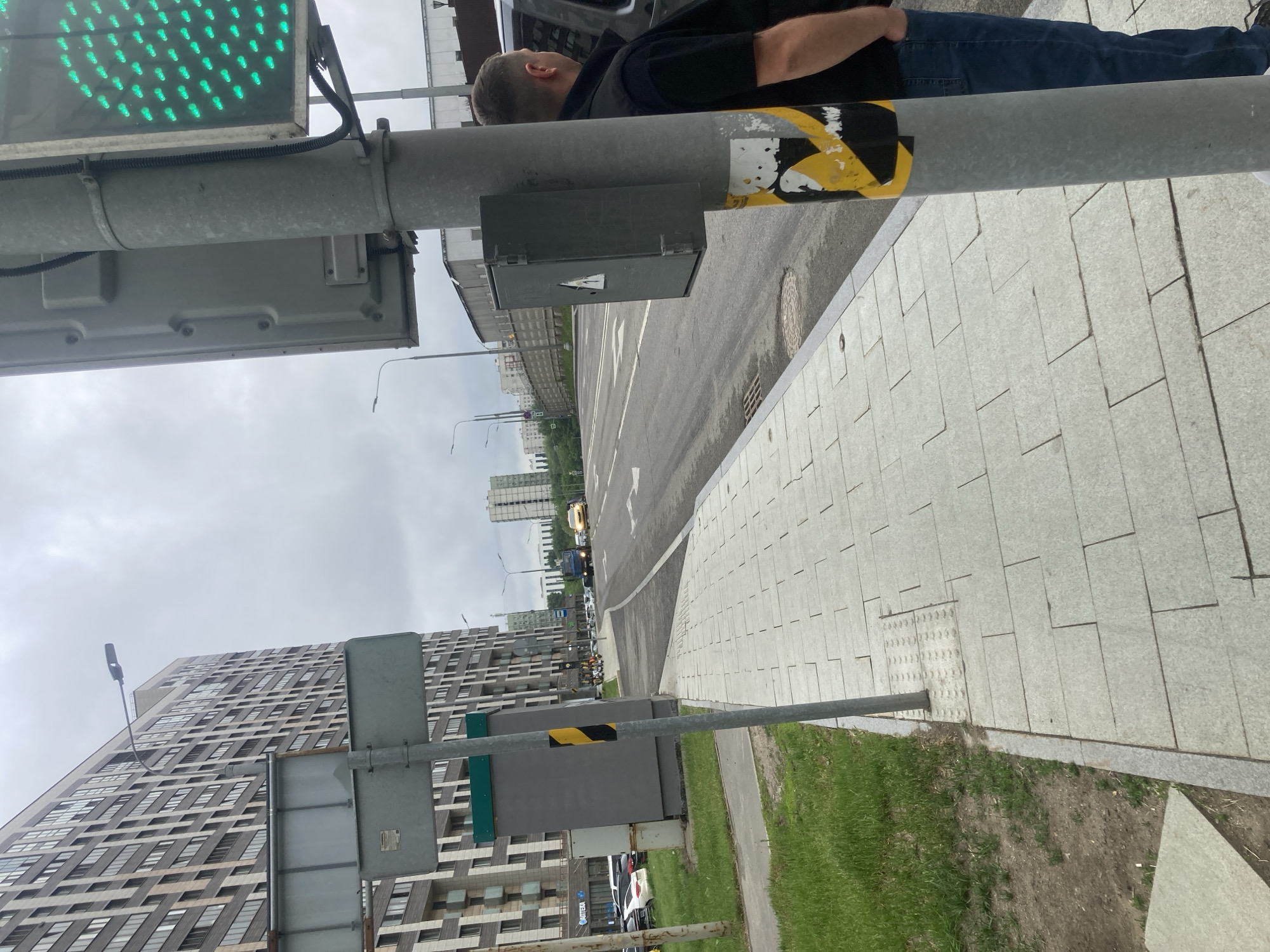 The image shows a street scene in Khimki, Russia. A person in jeans and a black shirt stands next to a traffic light pole, which displays a green light. The road is paved and there's a sidewalk made of light-colored square tiles adjacent to a grassy area. A tall building with many windows can be seen in the background. There are also other buildings and a few vehicles on the road. The sky is overcast.