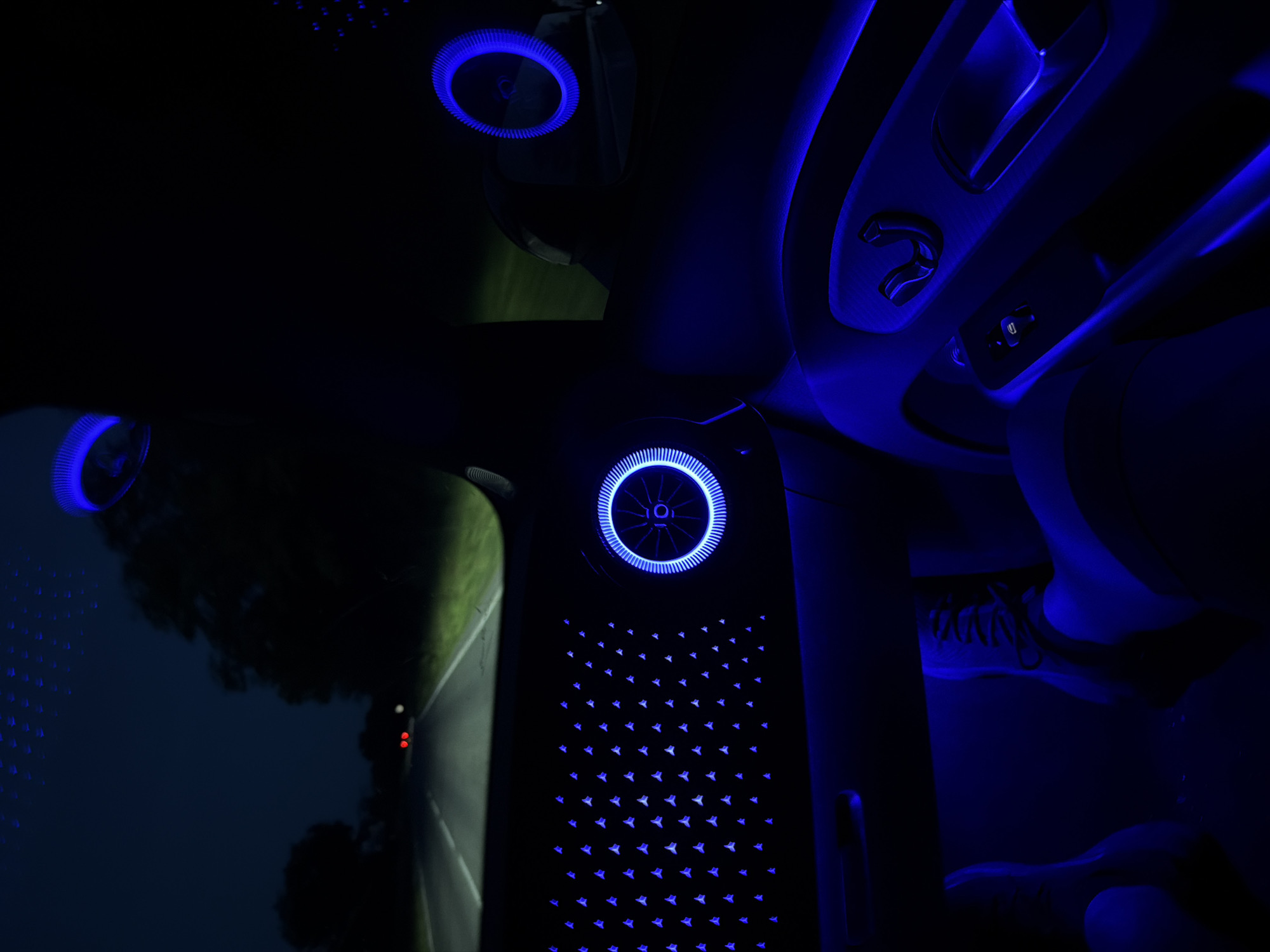 The image displays the interior of a vehicle at night, situated on a road in Obnova, Bulgaria. The car's cabin is extensively illuminated with blue ambient lighting.
The dashboard features a large panel adorned with a grid of numerous small, triangular blue lights. Adjacent to this panel on the right, a circular air vent is prominent, exhibiting a glowing blue LED ring and internal blue illumination. Similar blue light strips run along the lower sections of the dashboard and door panels on both sides of the vehicle. A second circular air vent, also featuring a blue LED ring, is visible in the upper right quadrant of the image, near the passenger side. The passenger door panel shows controls, likely for windows or seats, which are subtly lit by the blue ambient light. In the lower portion of the image, two feet are visible in the blue-illuminated footwell; one foot is shod in a dark sneaker with visible laces, indicating the presence of at least one occupant. Small blue light points are dispersed across the car's ceiling or reflected in the upper windshield.
Through the windshield, a dark asphalt road is visible, illuminated by the vehicle's headlights. White lane markings, including a solid line on the right and a broken line on the left, are discernible. Dark trees and vegetation line both sides of the road. In the distance, two distinct red lights, consistent with the taillights of another vehicle, are present. The external environment is dark, indicative of nighttime conditions.