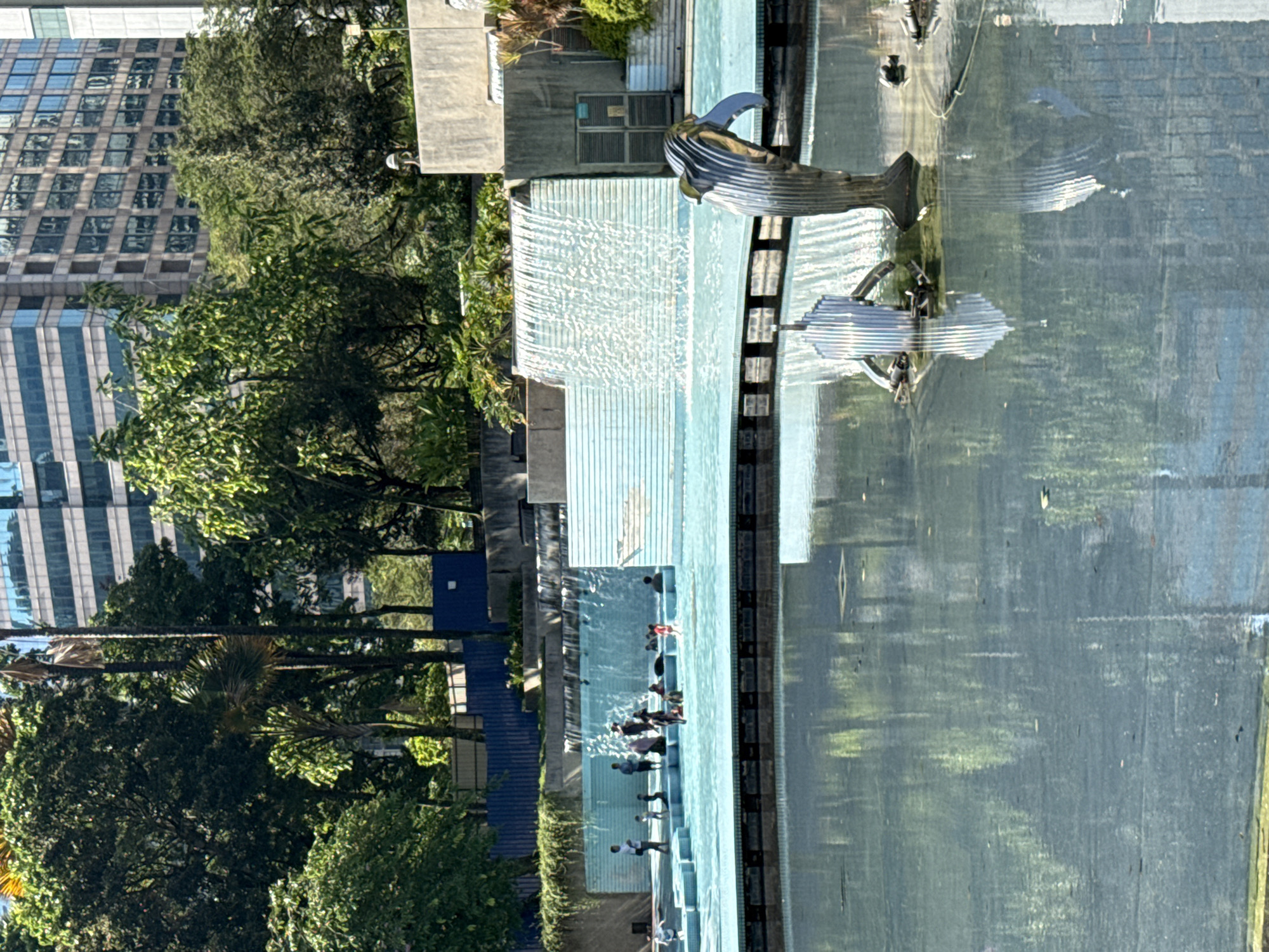 The image shows the Kuala Lumpur City Centre (KLCC) Park in Kuala Lumpur, Malaysia. The park features a large, tiered fountain with a sculpture of a whale. People are walking and standing near the fountain. There are trees and other greenery surrounding the fountain. Buildings are visible in the background.