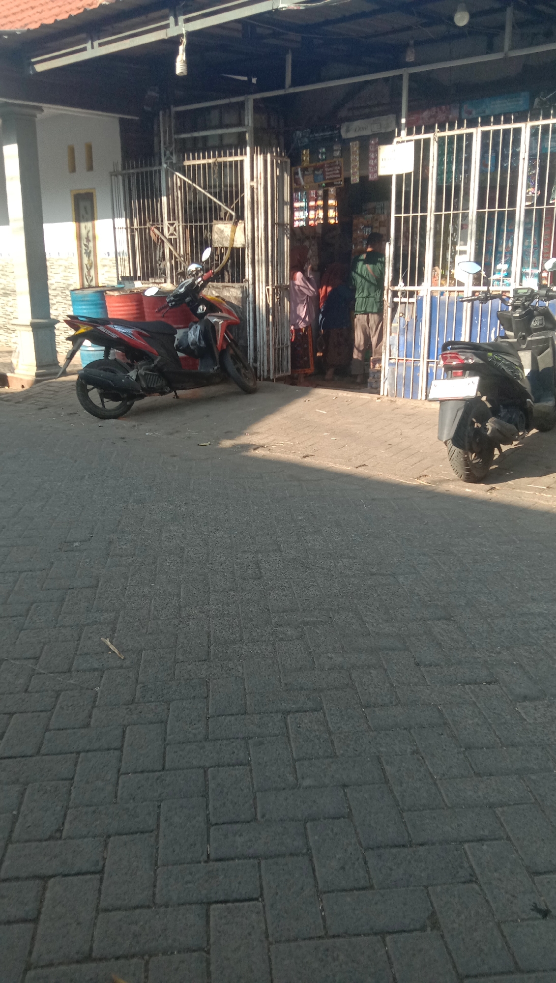 A street-level outdoor view captures a paved area in front of a shop in Karang Anyar, Indonesia. A red and black scooter is parked on the left, with its front wheel turned slightly to the left. Behind the scooter are two large metal barrels, one red and one blue. The shop, with a metal gate partially open, displays various items for sale. Three people are visible inside the shop: one wearing a light pink headscarf and a patterned skirt, another in a dark blue outfit, and a third in a green shirt. A dark-colored scooter is parked on the right side of the frame. The ground is covered with interlocking grey paving stones. The scene is bathed in sunlight, creating shadows on the ground.