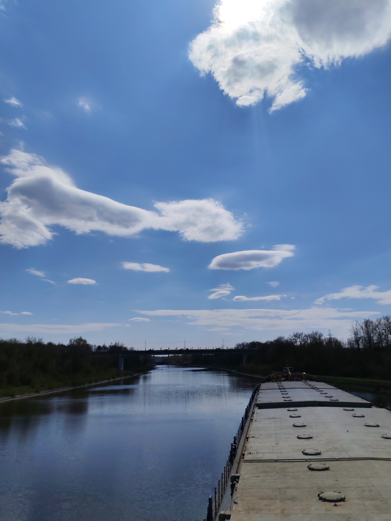 The image depicts a barge traveling on the Moscow Canal near the city of Yakhroma, Russia, under a bright blue sky dotted with scattered clouds. The canal stretches out in front of the barge towards a bridge in the distance, flanked by trees on either side. The water surface is reflective, mirroring the sky and surrounding vegetation. The deck of the barge occupies the foreground and right side of the frame, showing several hatches along its surface.