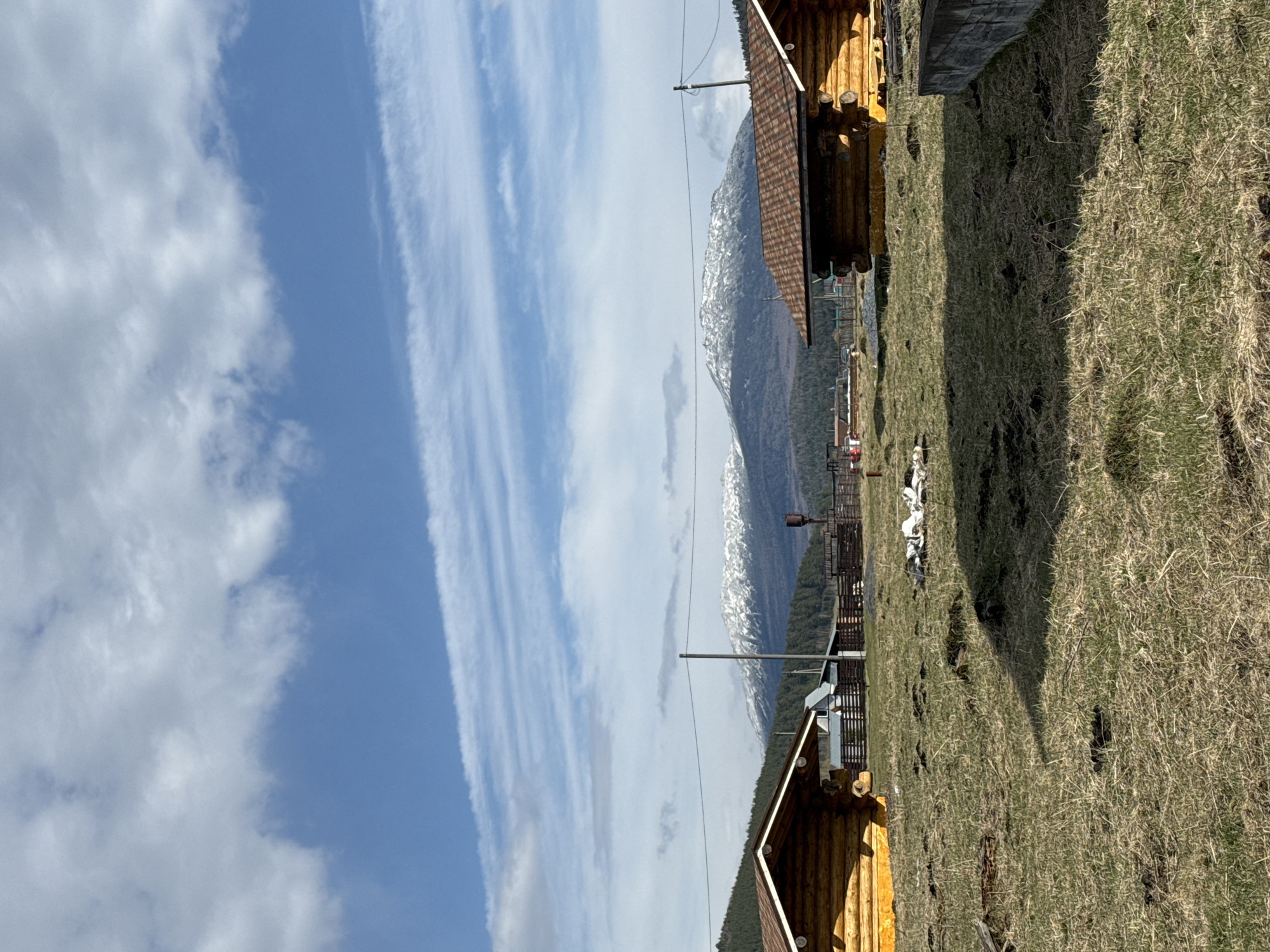 The image depicts a rural scene, possibly near the city of Tyulyuk, Russia, given the context. In the foreground are two wooden cabins with brown-tiled roofs, situated on a grassy slope. Patches of dry grass are visible. Beyond the cabins, a wire fence extends into the distance. A mountainous terrain with snow-capped peaks is visible in the midground. Above, the sky is filled with layered clouds and blue patches. Overhead power lines are strung from wooden poles. A small collection of rocks or debris is adjacent to a cabin.