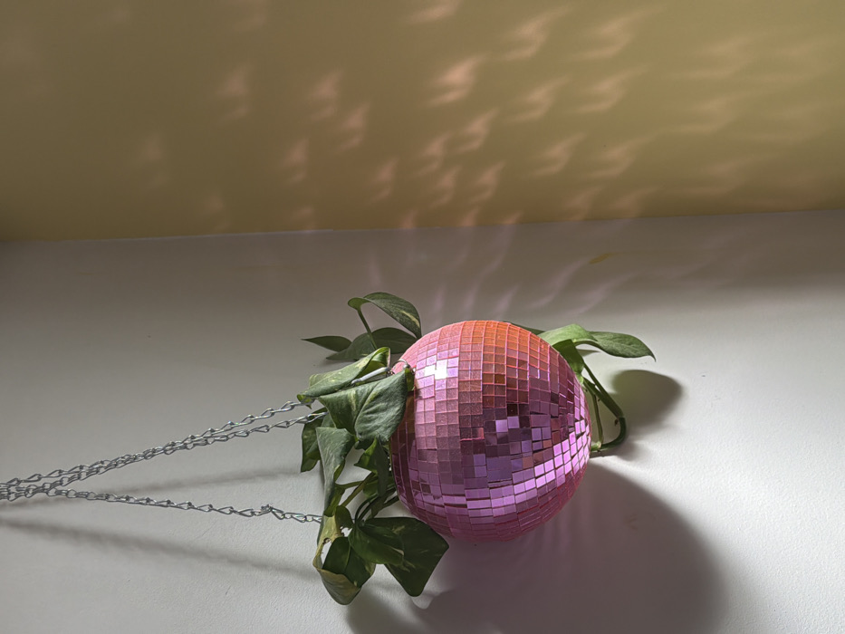The image depicts an interior room corner. Visual cues to identify a specific location within Tel-Aviv, Israel, are not present. A spherical hanging planter, constructed from numerous pink and some reddish-orange reflective mirror tiles, is suspended by three silver-colored metal chains from an unseen overhead point. The planter contains a live plant with multiple green leaves extending over the edges of the sphere, some exhibiting lighter green or variegated patterns. The plant casts a distinct shadow onto the white wall directly behind and beneath the planter. To the right, a second wall, colored light beige or pale yellow, meets the white wall at a perpendicular angle. Reflected light from the disco ball projects multiple small, bright, wavy, or crescent-shaped patterns onto the beige wall. No individuals are visible in the scene.