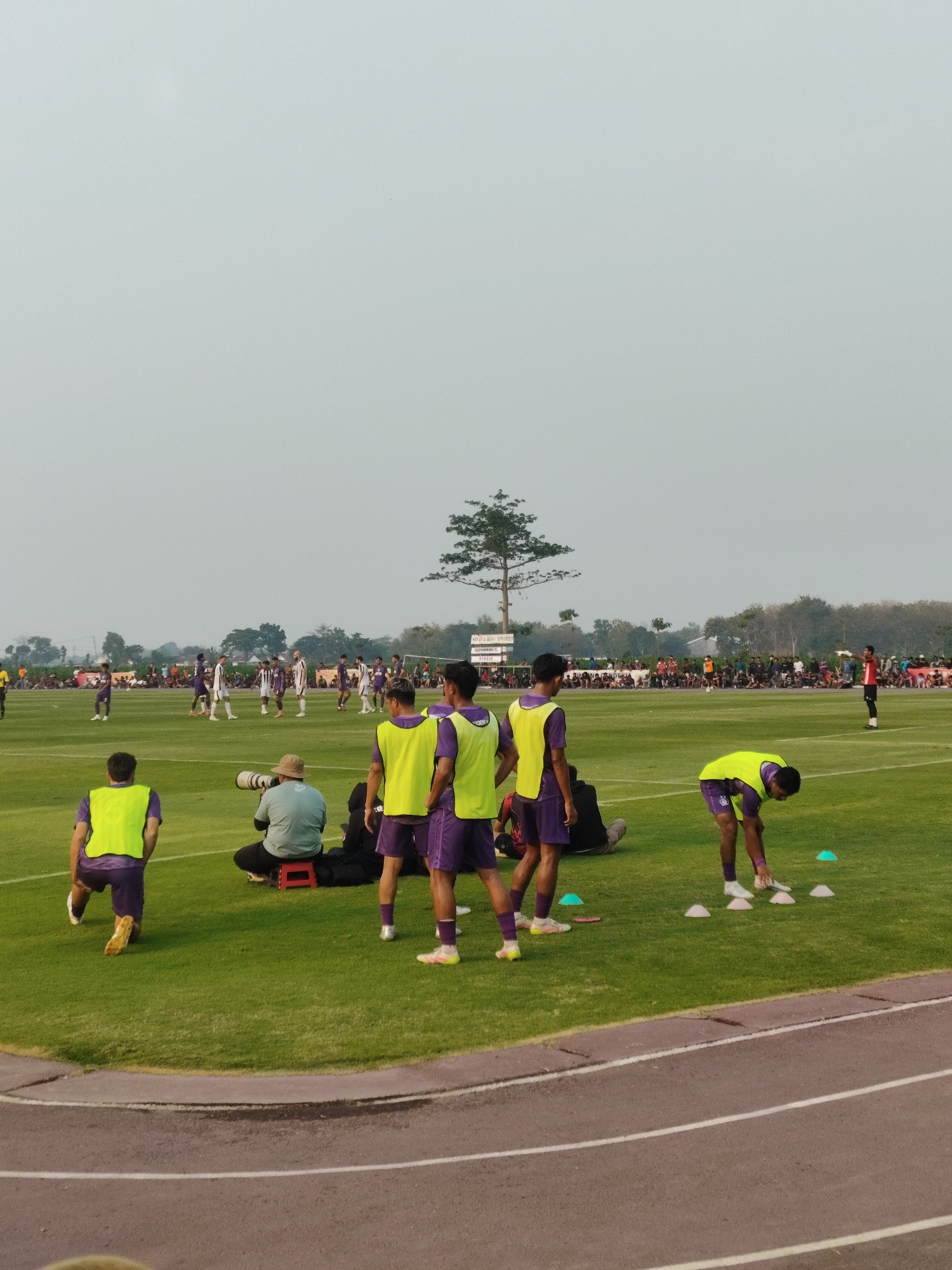 A group of young men in purple soccer uniforms and bright yellow vests are training on a grassy field at a stadium, likely in Batembat, Indonesia, as indicated by stadium signage. Some are stretching, while others are practicing drills around cones. A photographer with a large camera is seated on a red stool in the foreground, observing the players. Spectators are visible in the background, gathered along the edge of the field. The sky is overcast, and a single tree stands in the distance.