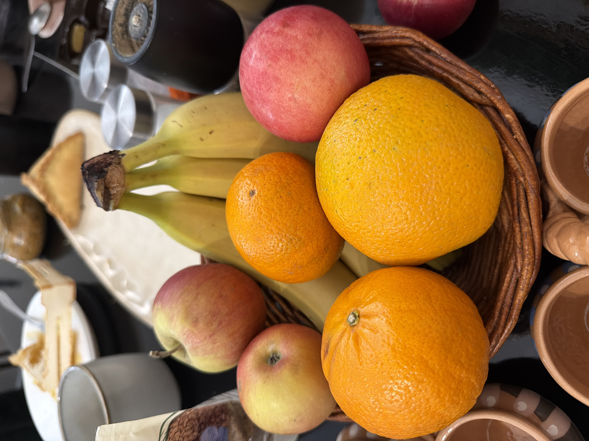 The image captures an overhead-angled view of a tabletop displaying a collection of food items. Central to the frame is a brown, woven wicker basket containing fresh fruit. Within the basket, a cluster of four to five yellow bananas with darkened stems is positioned towards the rear. Four oranges are visible: two prominent in the foreground, one brighter yellow-orange and one standard orange, with two more partially obscured behind. Three apples are present: two bi-colored red and yellow apples, and one predominantly red apple.
In the blurred background, on the left, a light grey ceramic mug and a white package with a floral design are visible. Further back, a white plate holds a partially consumed slice of a light-colored pastry or dessert, with a silver fork resting on it. To the right of this, an oval, light-colored dish contains a single triangular baked good. Beyond the fruit basket, a black cylindrical object, likely a grinder, stands next to two silver-topped shakers.
In the foreground, along the bottom edge, several small, unglazed reddish-brown ceramic pots or cups are arranged, one featuring a white dotted pattern. The items are situated on a dark, reflective surface, indicative of a table. The scene is located in Pervomaysky, Russia. No individuals are present, and no active interactions are depicted.
