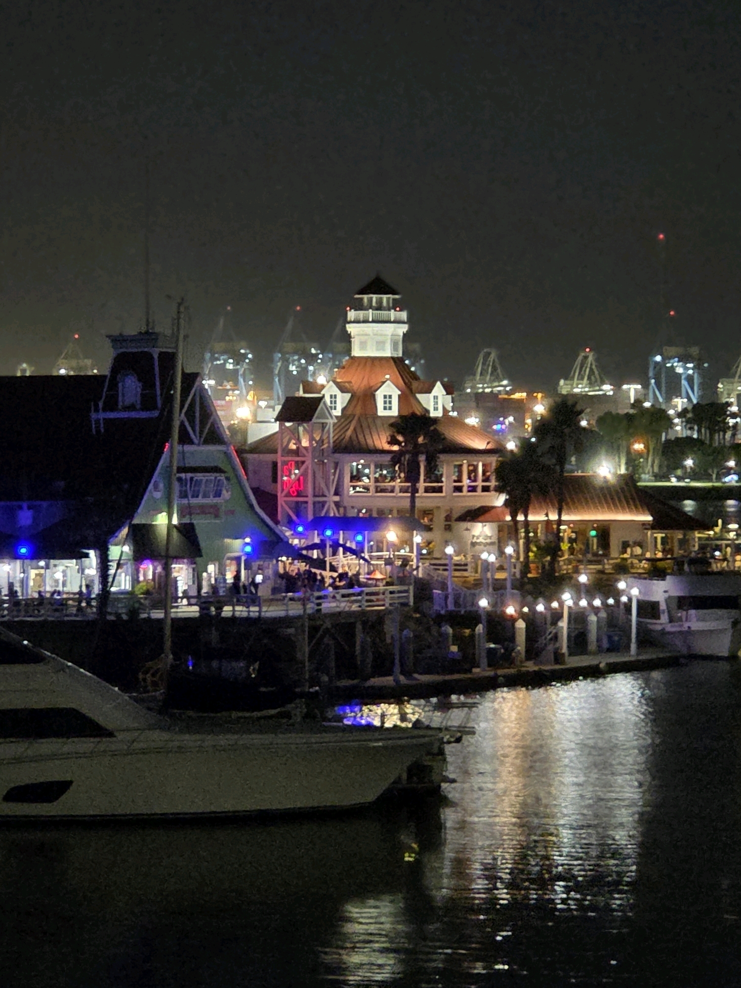 The image depicts the nighttime view of the waterfront area in Long Beach, California, USA. Several buildings with illuminated windows and brightly lit decorative lights are visible. A prominent structure with a red roof and a white tower stands out. Boats are docked along the waterfront, with one large white yacht prominently positioned in the foreground. The water reflects the lights from the buildings and boats, creating a shimmering effect. In the background, city lights and distant structures, possibly including amusement park rides and cranes, are faintly visible against the dark night sky. The overall scene conveys a lively and illuminated waterfront ambiance.