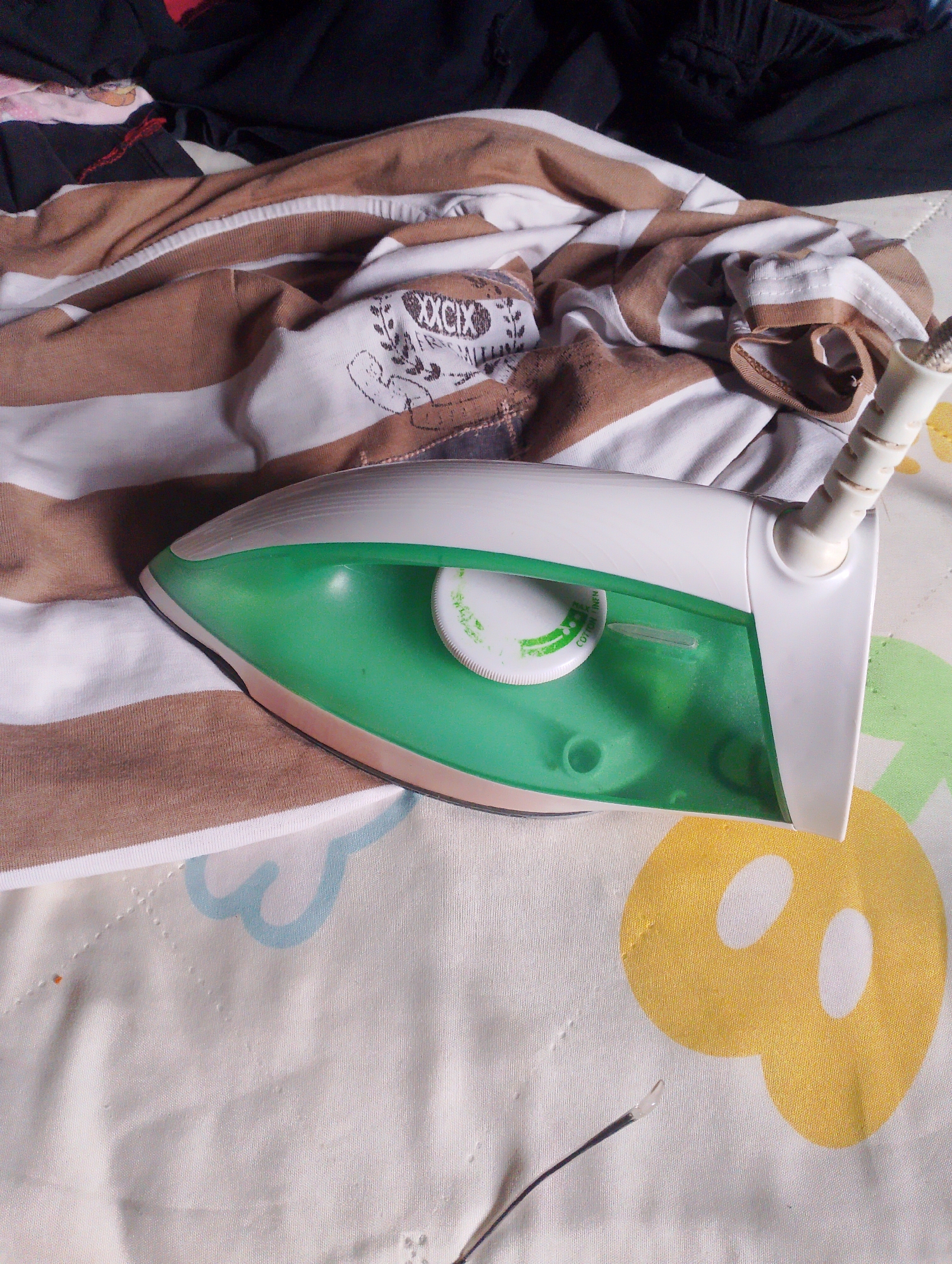 An iron is placed on a striped shirt, part of a laundry pile. The iron is green and white, with a transparent water reservoir. The shirt is brown and white striped with a graphic design on it. The laundry is on a white surface with yellow and blue patterns. The image is taken from a high angle, focusing on the iron and shirt. The scene appears to be indoors, likely a home, and does not provide any specific information about the city of Panyubrangan, Indonesia.