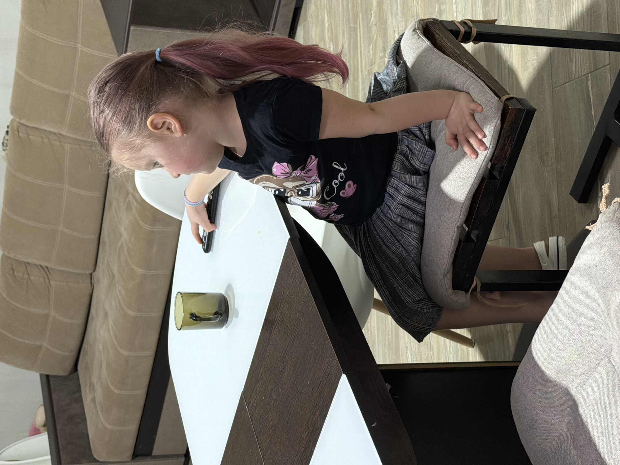 This indoor image shows a young girl seated at a white table in what appears to be a living room or dining area in Chaikovsky, Russia. The girl has light brown hair with pink highlights, tied back in a ponytail with a light blue elastic. She wears a black short-sleeved t-shirt featuring a graphic of a cartoon animal with a pink bow and the word "Cool," along with a plaid skirt, and white sneakers with black stripes. A colorful bracelet is visible on her left wrist.
She is intently looking down at a black remote control or small electronic device held in her left hand, which rests on the white tabletop. To her right on the table is a dark green glass mug or cup, possibly with a suction cup base.
The background features a light beige or tan sectional sofa. The floor appears to be made of light-colored wooden planks. The girl is sitting in a wooden chair with light grey cushions. The overall setting suggests a casual, domestic environment. The lighting is ambient, indicating an indoor shot without strong directional sunlight. The girl's expression conveys focus or concentration on the object in her hand.