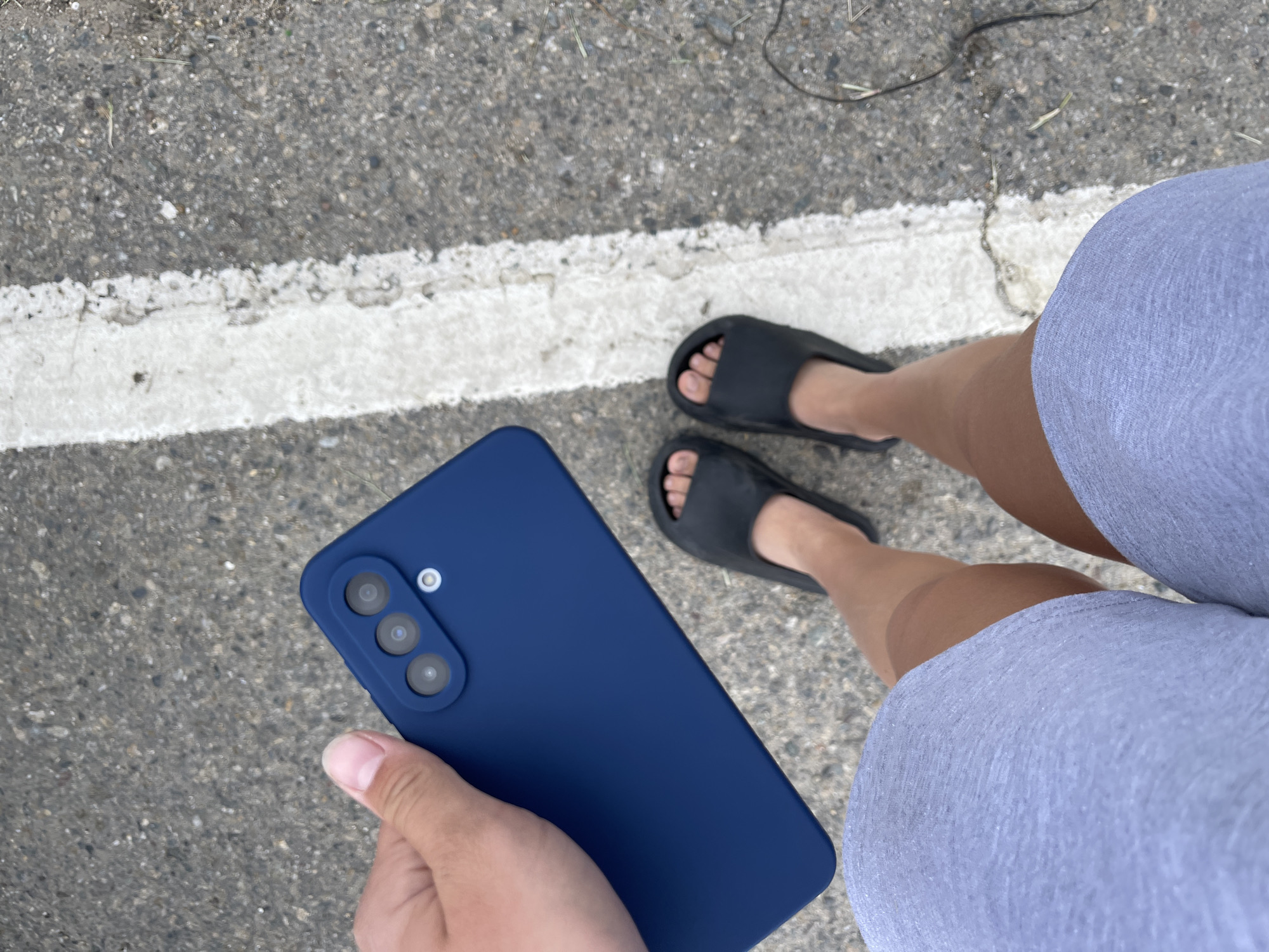 A person is holding a dark blue smartphone in their left hand, positioned over their right thigh which is covered by a light grey pair of shorts. The phone has a triple camera module. The person is wearing black slide sandals on both feet. They are standing on an asphalt surface with a white painted line running vertically along the right side of the frame. The background is a grey asphalt surface with scattered debris and small green blades of grass. The image is taken from a high-angle perspective, looking down at the person's legs and the phone. The lighting suggests it is daytime. The scene appears to be outdoors, possibly on a street or sidewalk in Moskovskoe, Russia.