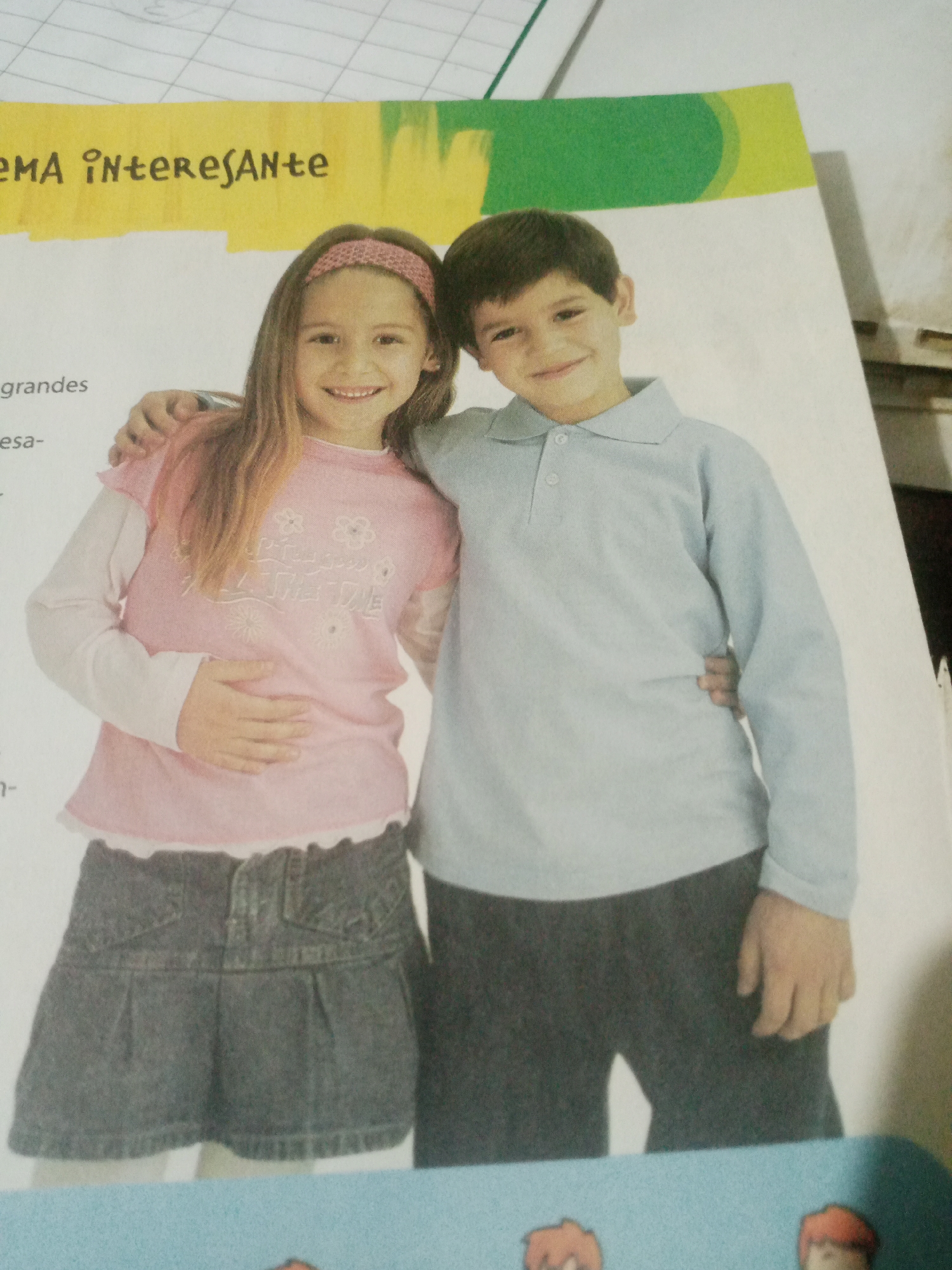 The image shows a young boy and a young girl, both smiling, with their arms around each other. They are standing in front of a page from what appears to be a book or magazine, with some text visible. The girl is wearing a pink shirt with white trim and a denim skirt. The boy is wearing a light blue long-sleeved shirt and dark pants. The background is a white page with some printed text. No specific location in Santa Ana, Venezuela can be determined from the image.