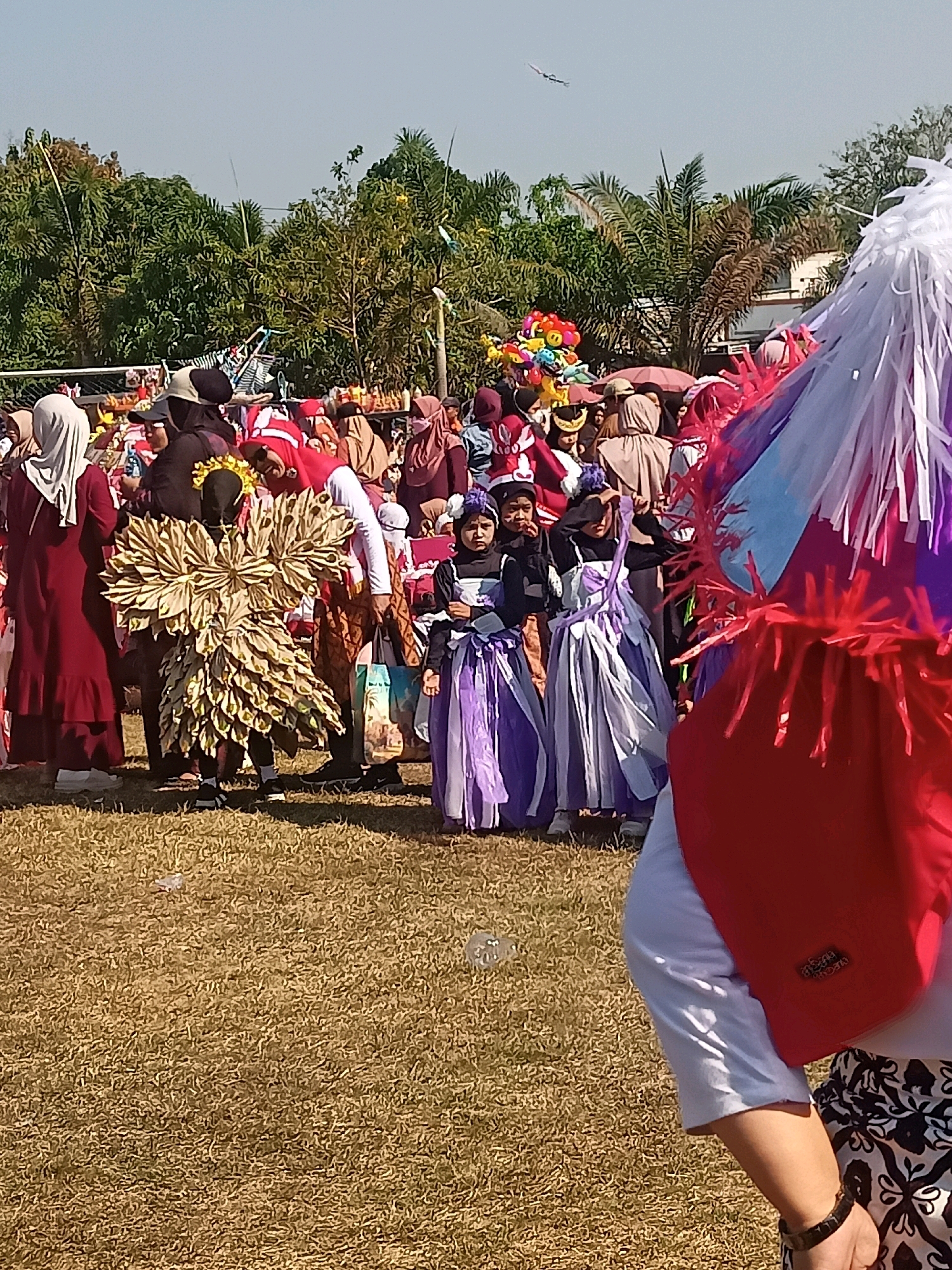 The image depicts an outdoor gathering of people, possibly a festival or parade, in Sumberlawang, Indonesia. The scene is predominantly grassy with trees in the background. Several individuals are dressed in elaborate costumes. In the foreground, a person is partially visible from behind, wearing a red backpack and a patterned skirt. In the middle ground, a child stands out in a large, layered, golden-yellow costume resembling wings or petals. Other children are also present, some in purple and white striped dresses. A crowd of adults, many wearing hijabs and traditional clothing, are visible behind the costumed children. A cluster of colorful balloons is held by someone in the crowd. The lighting suggests it is a sunny day.