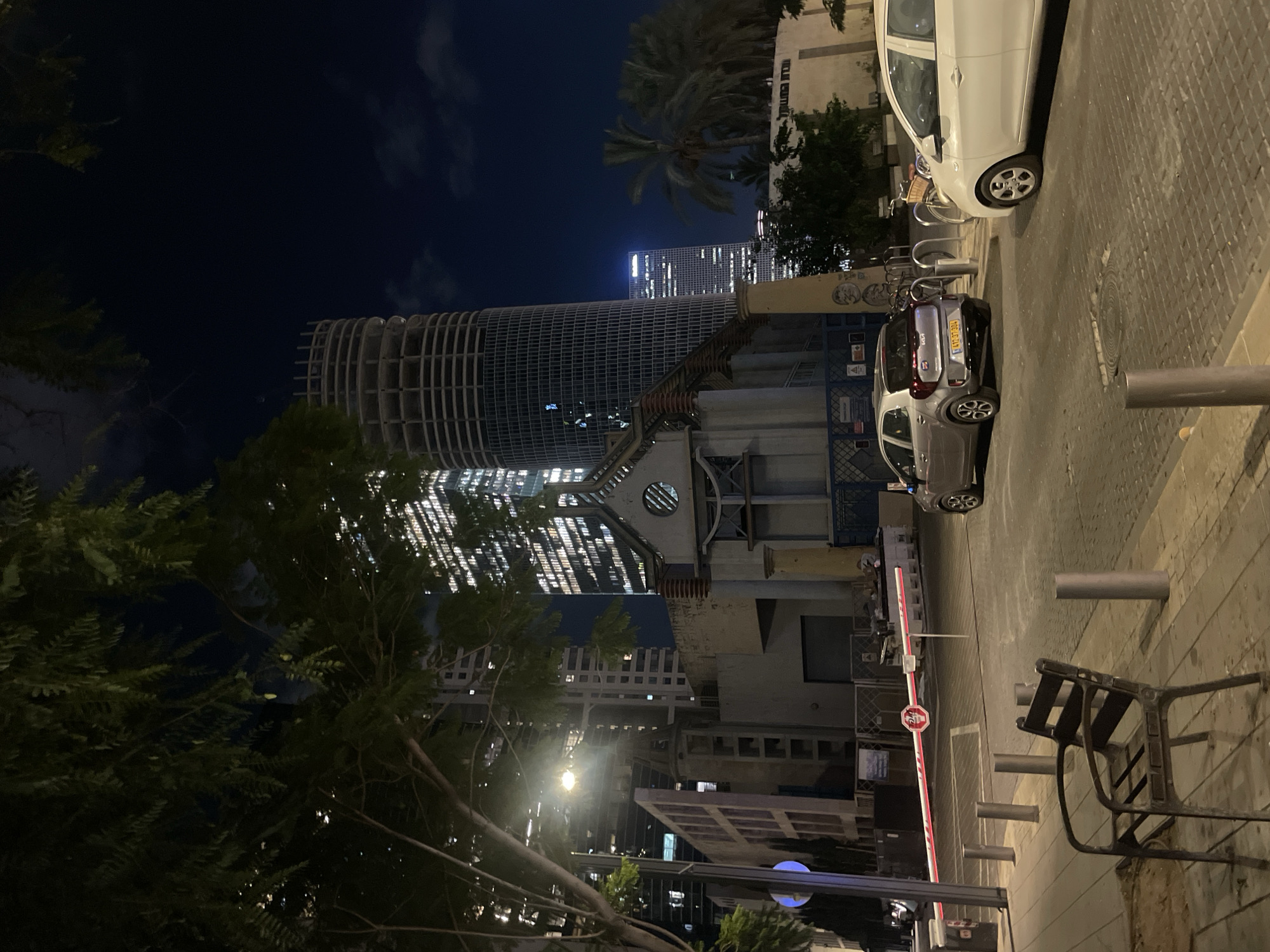 The image depicts an urban street scene at night in Tel Aviv, Israel, likely in a business district given the modern skyscrapers. In the foreground, a dark metal chair sits on a paved sidewalk. A red and white striped barrier arm with a "STOP" sign is lowered across the street, indicating a checkpoint or entrance. Two cars are visible on the street: a silver Kia Picanto on the left and a white car on the right, partially obscured. The background features several tall, illuminated buildings, including a distinctive circular, unfinished skyscraper under construction. A smaller, older building with an arched window is situated in front of the skyscrapers. Palm trees are visible to the right, suggesting a warm climate. The overall atmosphere is one of a quiet, nighttime city street with ongoing development.