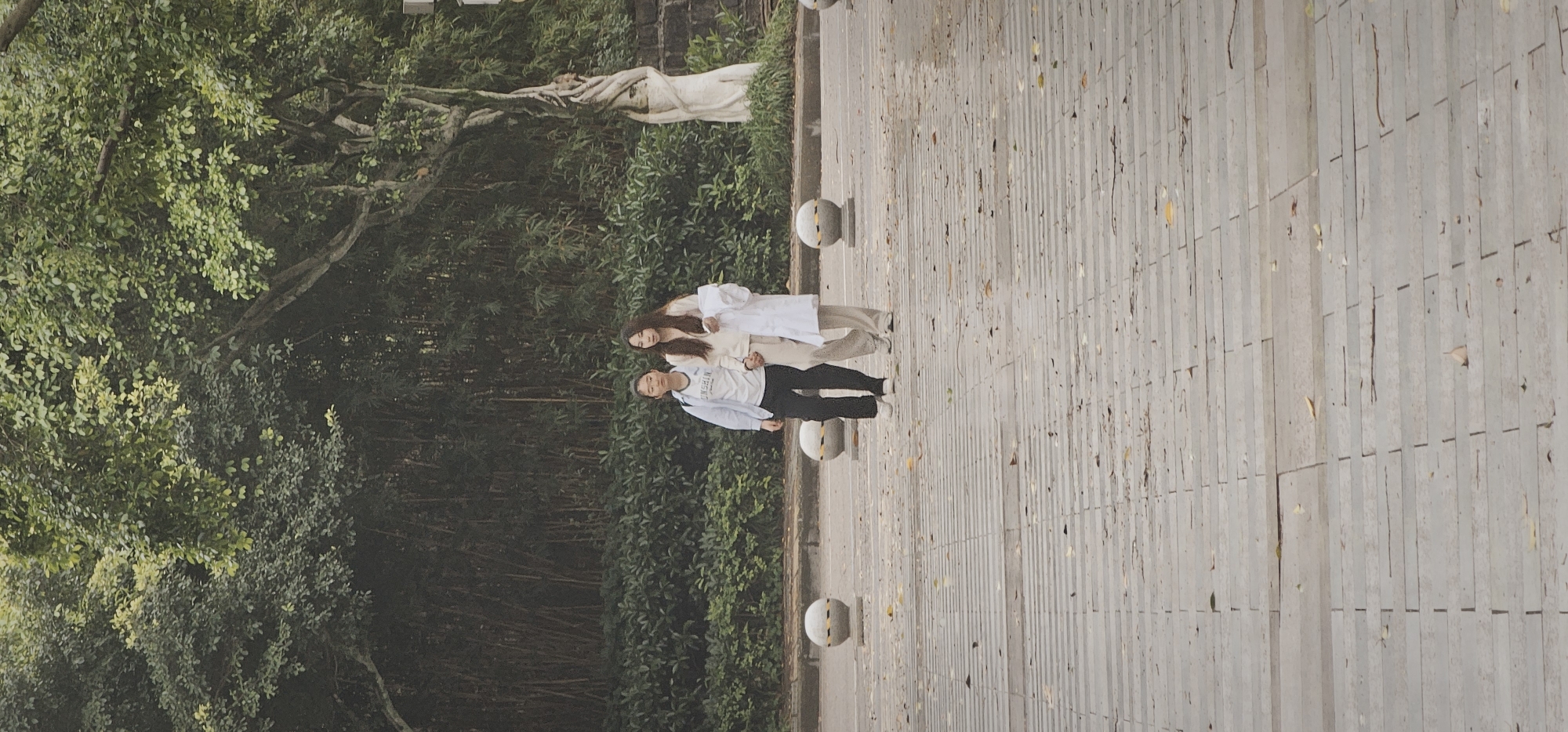 A couple stands on a stone-tiled walkway in the Chongqing High-tech Zone, China. The man wears a white sweatshirt, blue button-up shirt, and black pants, while the woman has on a white top and light tan pants with a white jacket. They are positioned beside a dense, green forest. The walkway is bordered by stone spheres. A statue is partially visible in the background, adjacent to the trees.