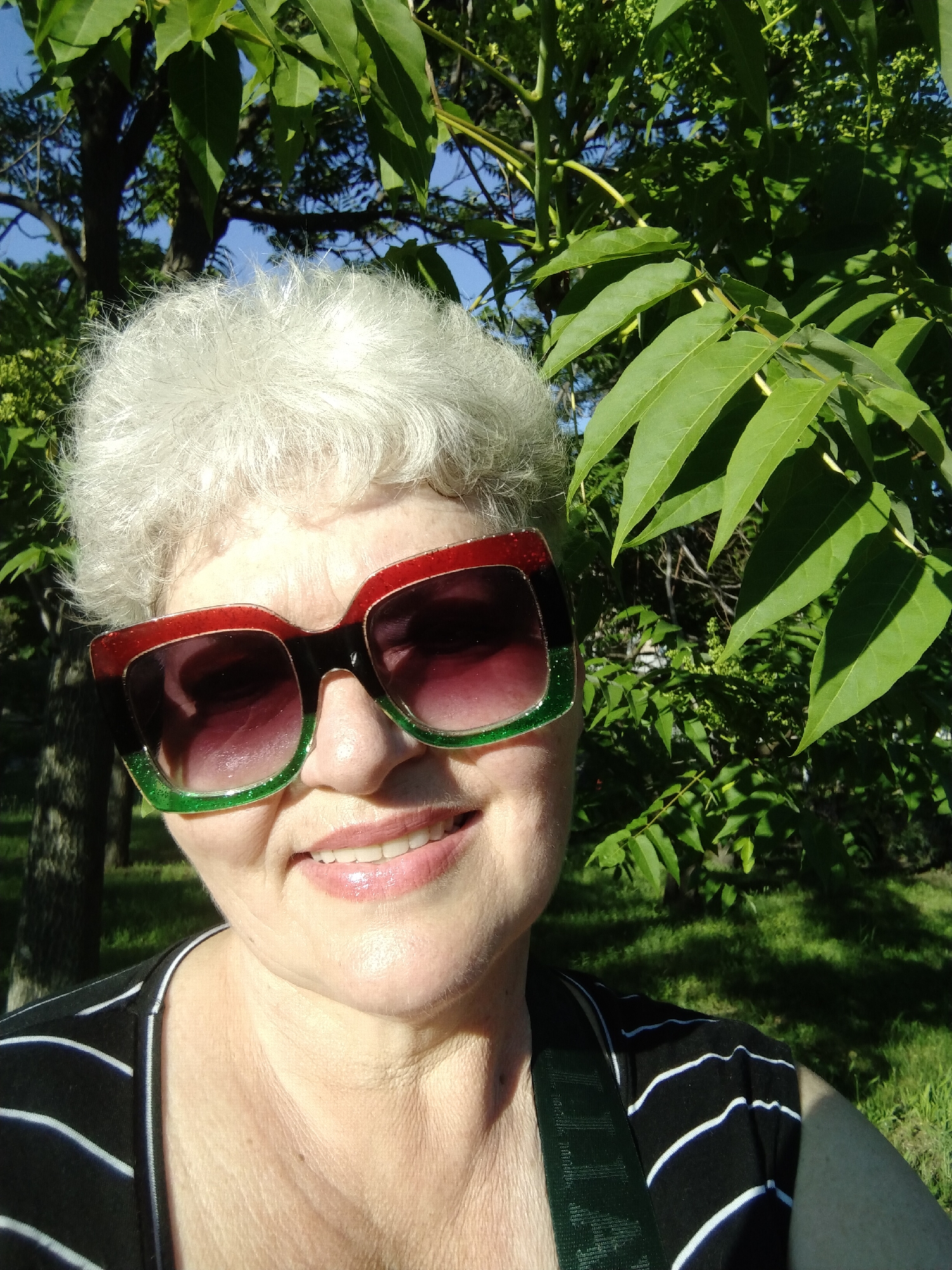 A close-up, eye-level selfie depicts a smiling woman outdoors in Horishni Plavni, Ukraine. The woman has short, curly, white hair and is wearing oversized, square-framed sunglasses that are glittery red on the upper half and glittery green on the lower half, separated by a black line across the center. Her lips are parted in a smile, revealing teeth and covered in a pink gloss. She wears a black top with thin white horizontal stripes, and a dark green strap is visible over her right shoulder. The background features dense green foliage from trees and shrubs, some displaying small green clusters, with portions of clear blue sky visible through the leaves. Green grass covers the ground in the lower right, partially shadowed. The scene is brightly illuminated by natural sunlight.