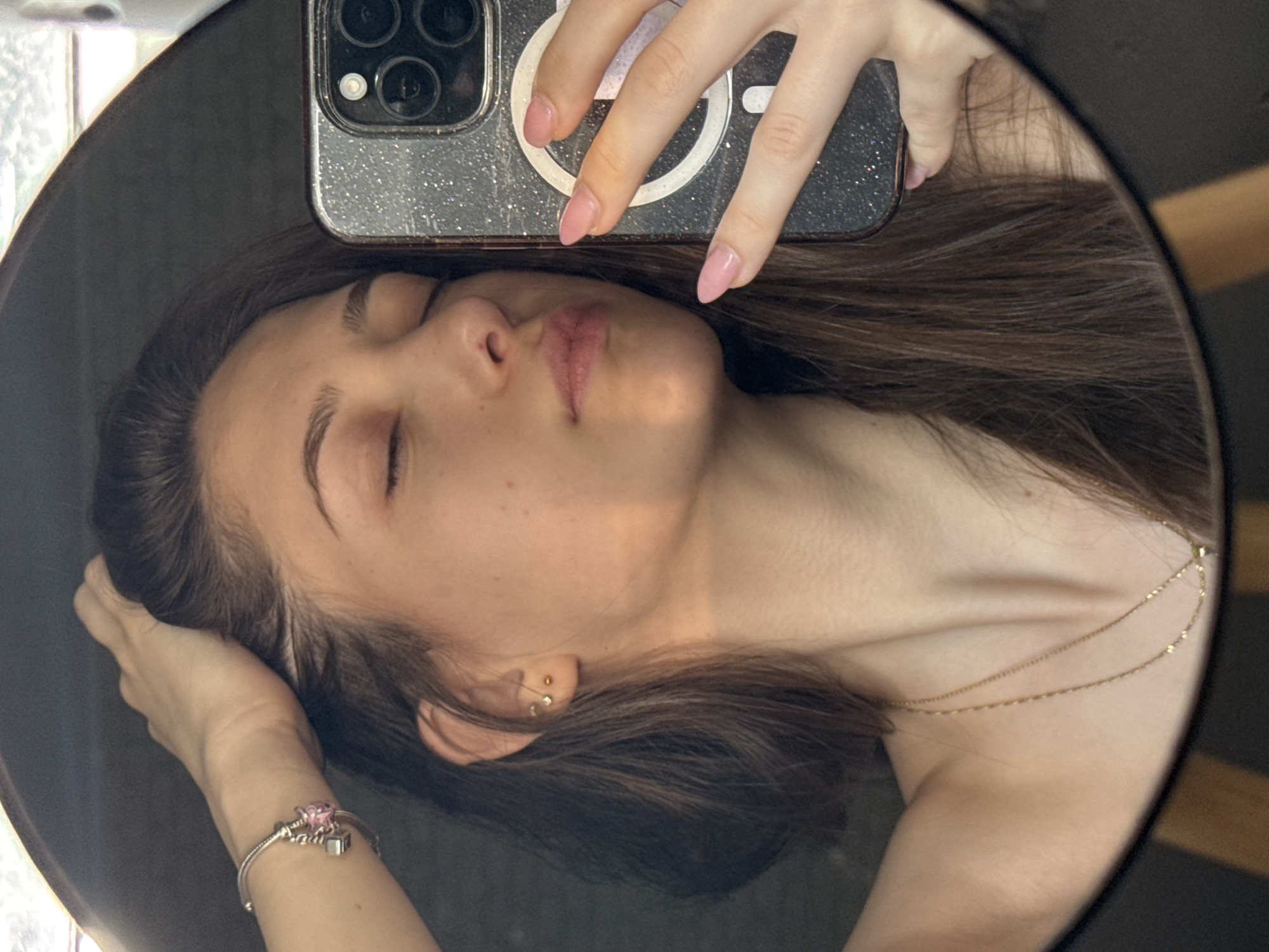 The image is a selfie taken in a mirror. A fair-skinned young woman is lying down, facing the camera, eyes closed. She is holding a mobile phone, a silver iPhone, to take the picture. Her hair is brown and straight. She is wearing a thin gold necklace and a bracelet on her left wrist. Her nails are painted pale pink. There are no indicators of location in the city of Sofia, Bulgaria.