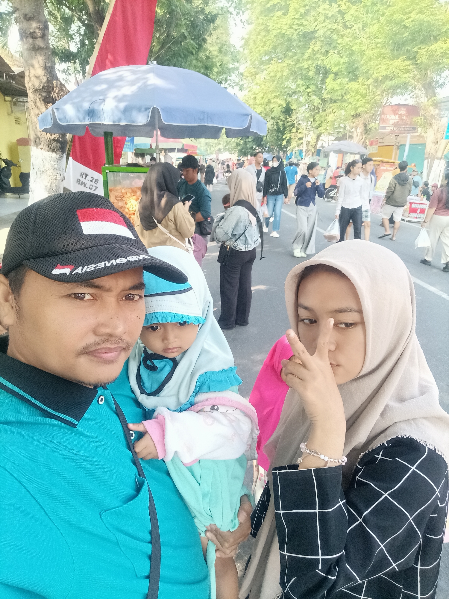 A family of three, a man, a woman, and a baby, are posing for a photo in front of a street market in Pare, Indonesia. The man in the foreground is wearing a turquoise polo shirt and a black baseball cap with the Indonesian flag on it. He is holding the baby, who is wearing a light blue hijab and outfit. To the right of the man, a young woman in a beige hijab is making a peace sign with her fingers. In the background, a crowd of people are browsing various stalls and food vendors, some of which are under umbrellas. Trees line the street, and the sky is bright and sunny. The overall scene depicts a lively outdoor market environment in a city.