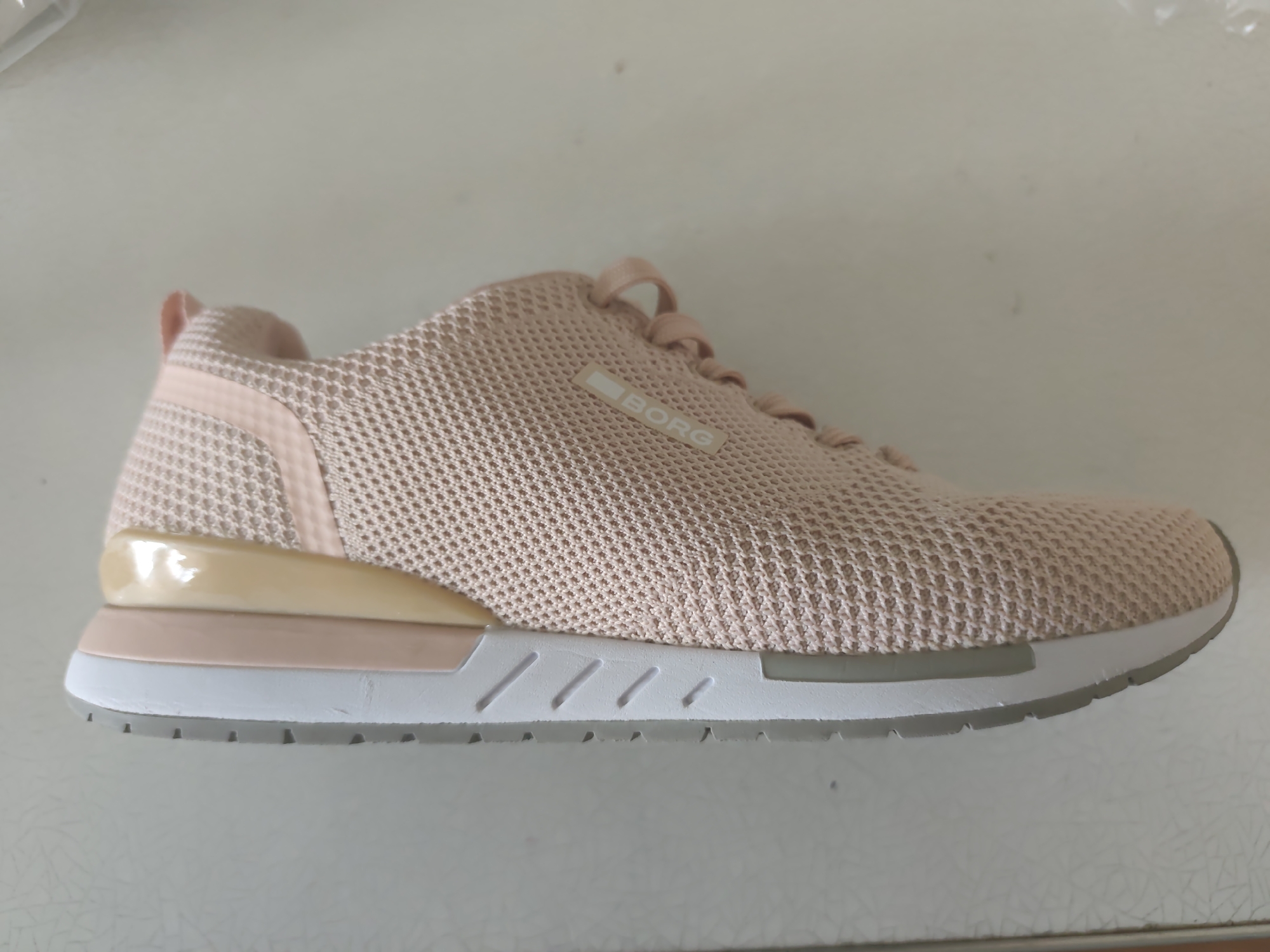 A single, light pink mesh sneaker with a light tan and white sole is displayed in a side profile view. The sneaker is positioned horizontally on a light-colored, textured surface, likely a table or countertop. The sole has a layered appearance, with a white midsole and a gray outsole featuring a pattern of small, rectangular indentations. A small, rectangular logo with the word "BORG" is visible on the side of the sneaker's upper. The background is a plain, light gray surface, suggesting an indoor setting, possibly a retail display or a personal collection. There is no indication of the specific location within the city of Kyiv, Ukraine.
